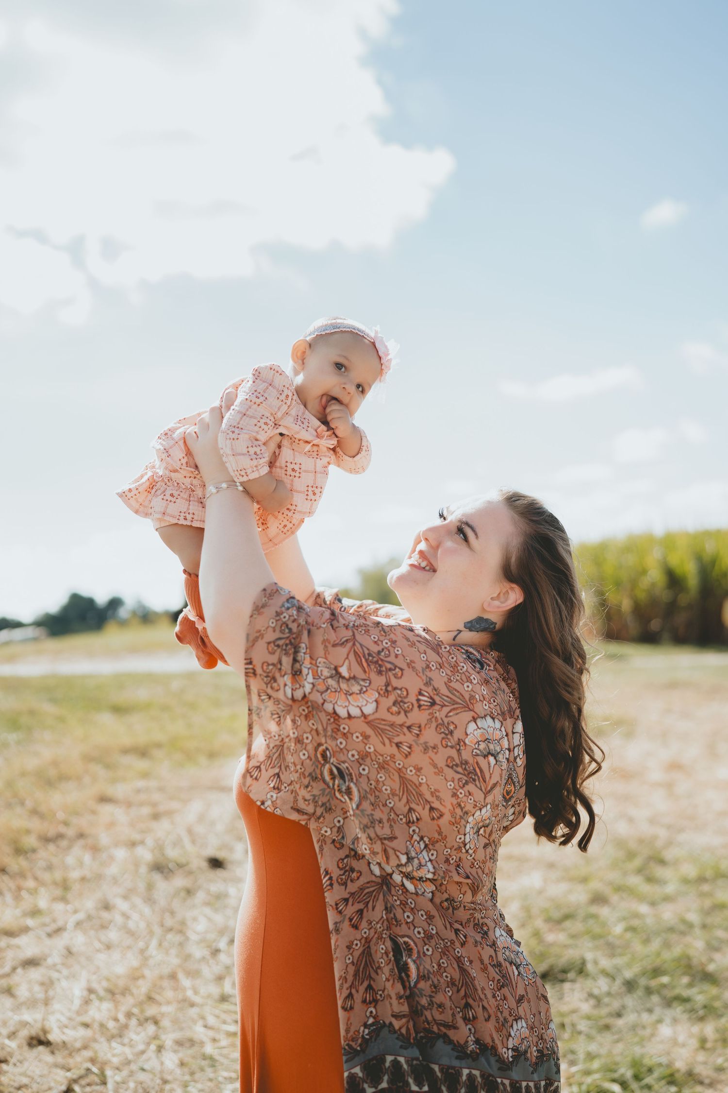 Smiling mother lifting happy baby in pink dress outdoors on a sunny day.