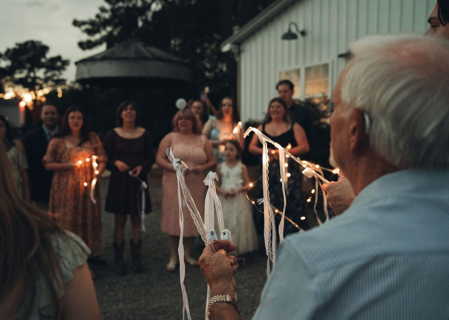 Wedding guests hold sparklers during an evening outdoor celebration near a rustic barn venue.