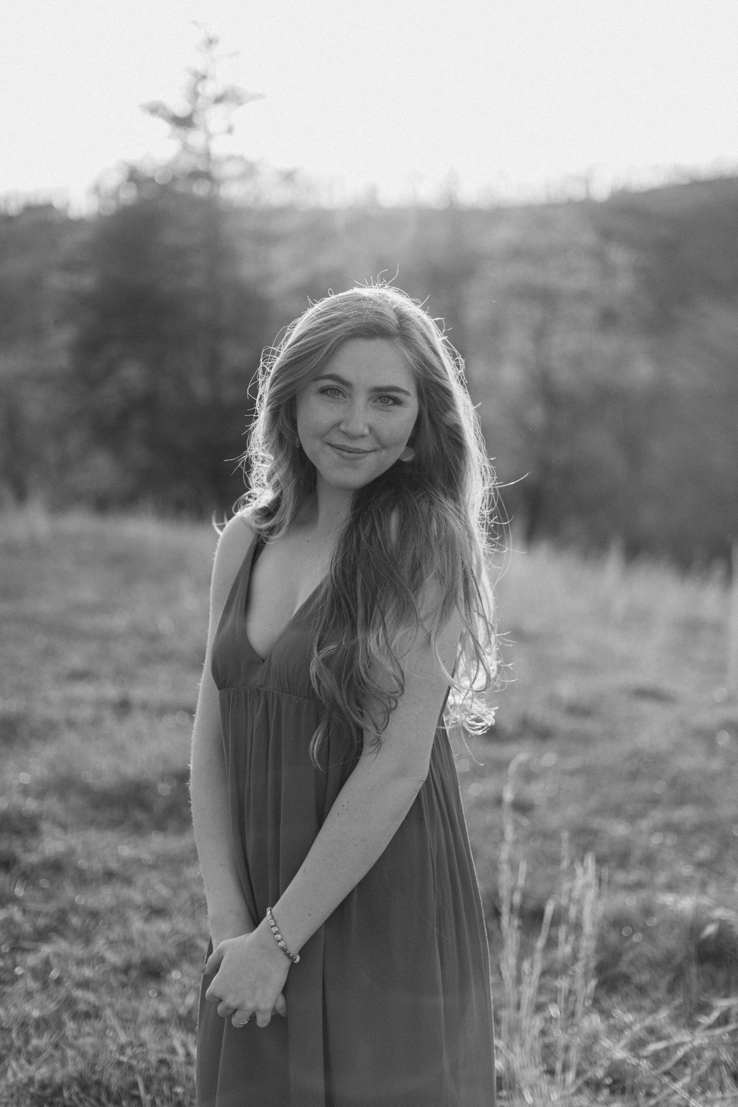 Black and white portrait of a young woman with long wavy hair in a sundress standing in a natural outdoor field.