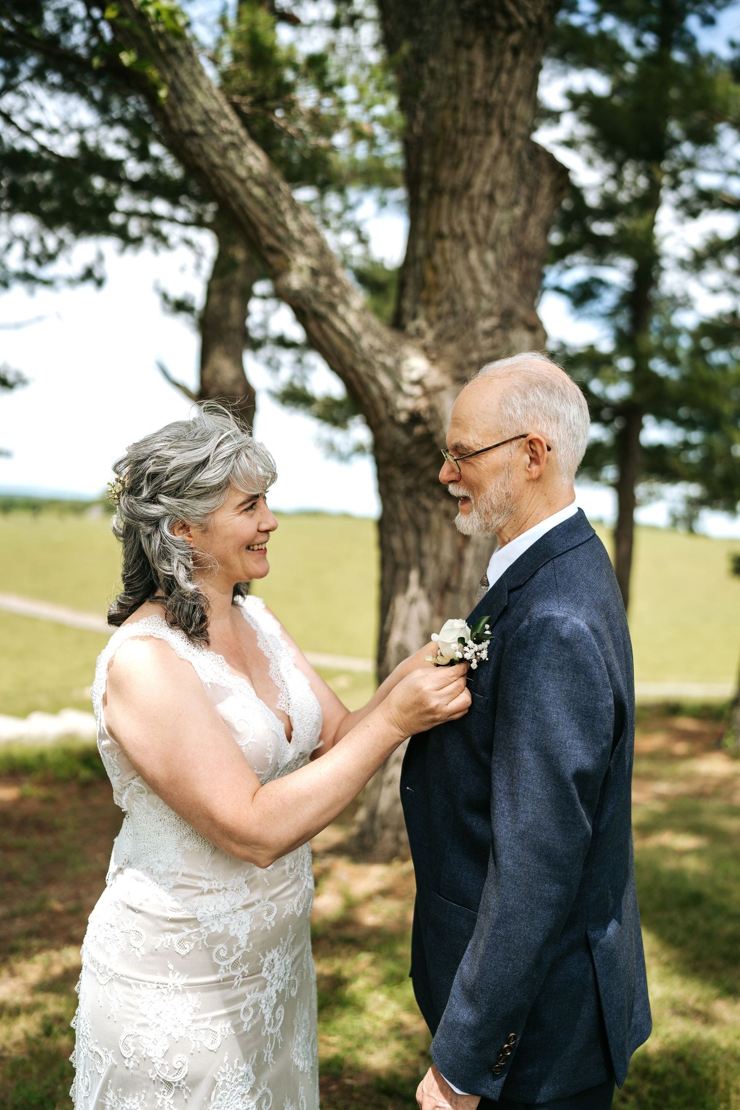 Smiling mature bride adjusts boutonniere on groom in navy suit during outdoor first look moment.