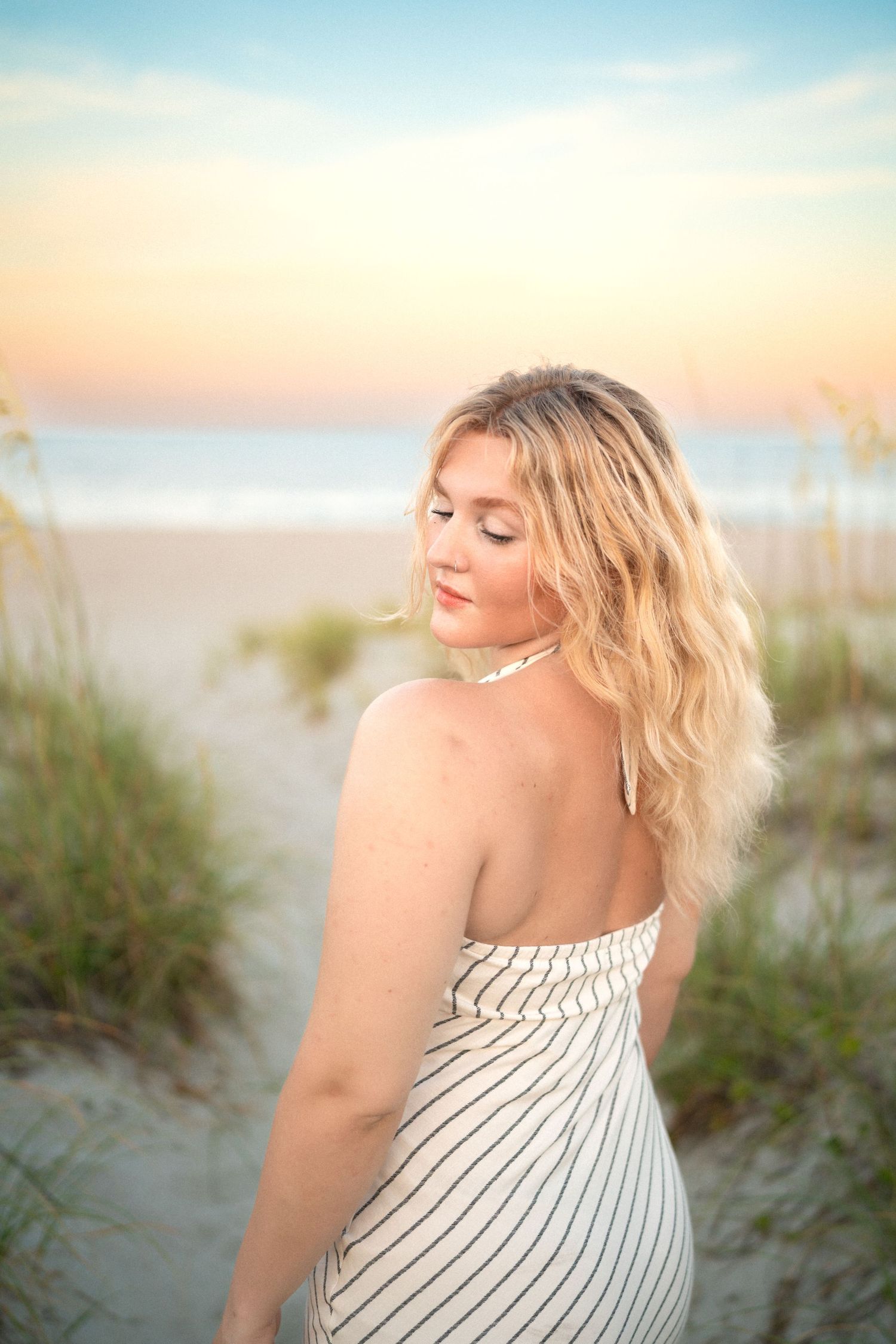 Blonde woman in striped halter dress posing on sandy beach dunes at golden hour sunset.