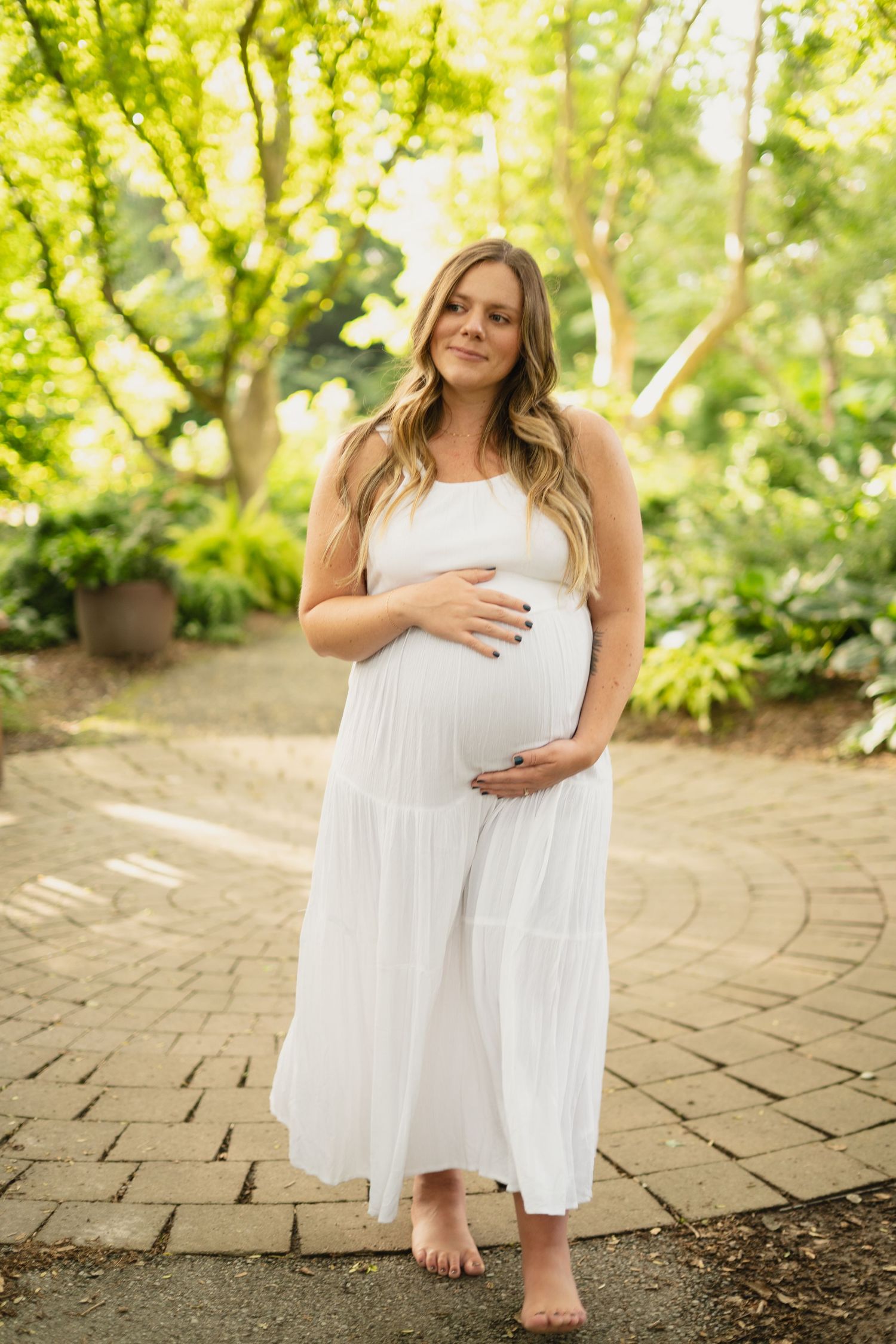 Pregnant woman in white maxi dress cradling baby bump in sunny garden.