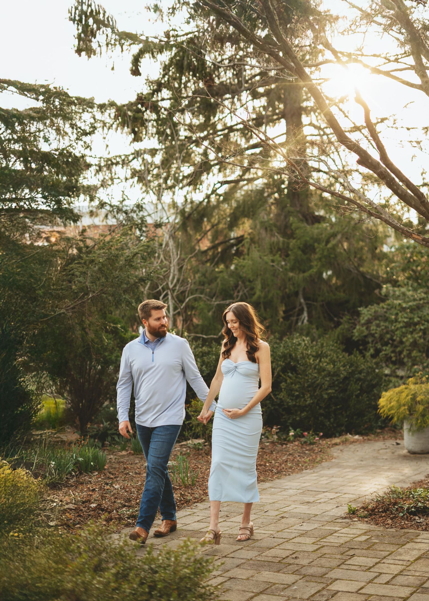 Pregnant couple walking hand in hand on a sunlit garden path during a beautiful outdoor maternity photo session.