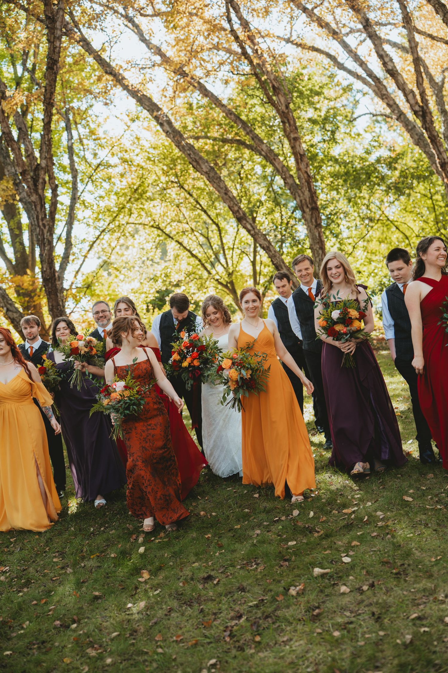 Wedding party walking outdoors in fall-colored mismatched gowns of yellow, orange, and burgundy amid autumn trees.