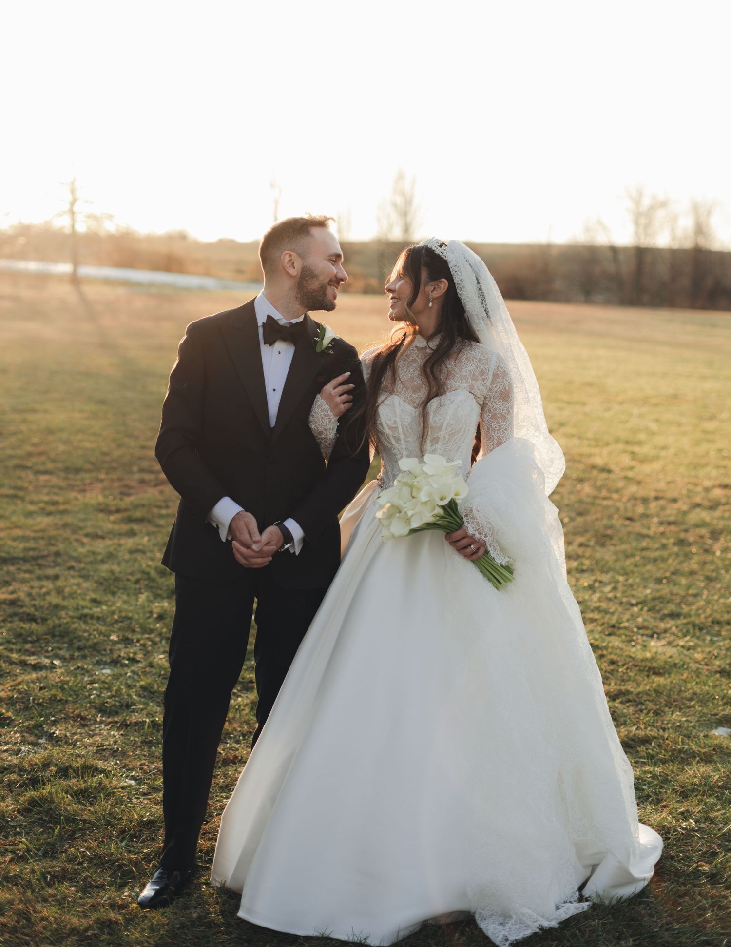 Elegant bride and groom walking hand in hand across a golden field at sunset, exchanging loving glances.