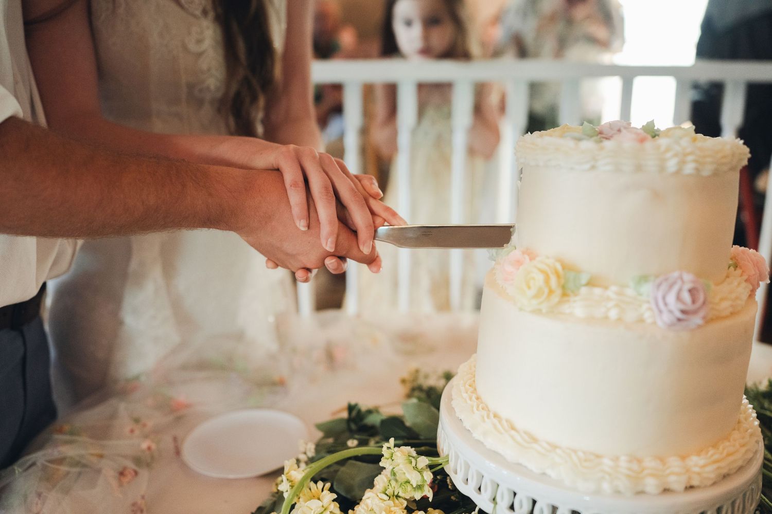 Bride and groom cutting elegant two-tier white wedding cake decorated with pastel flowers.
