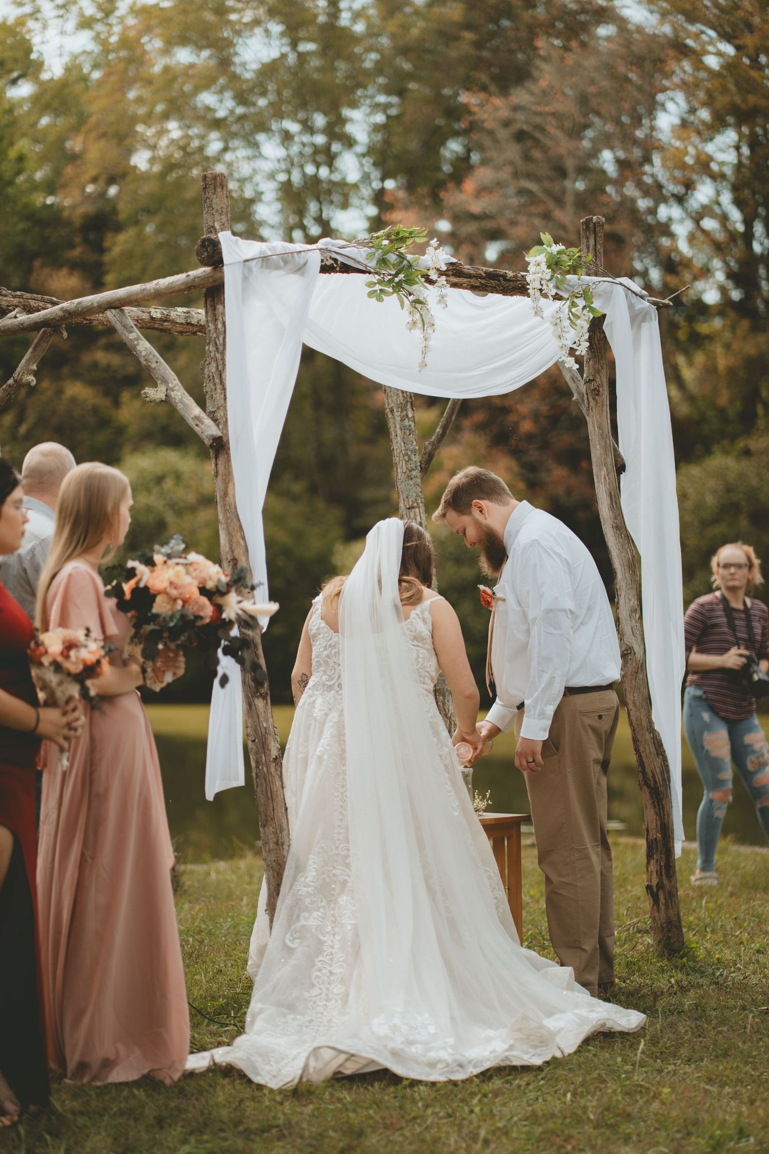 Bride and groom hold hands under rustic branch wedding arch with white draping and floral accents.