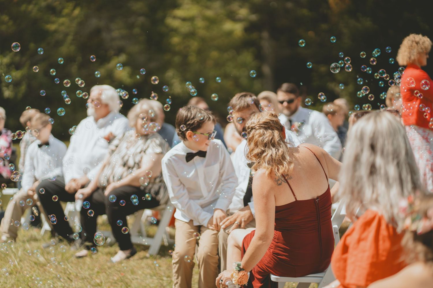 Young boy in bow tie watches bubbles float through outdoor wedding ceremony with seated guests.