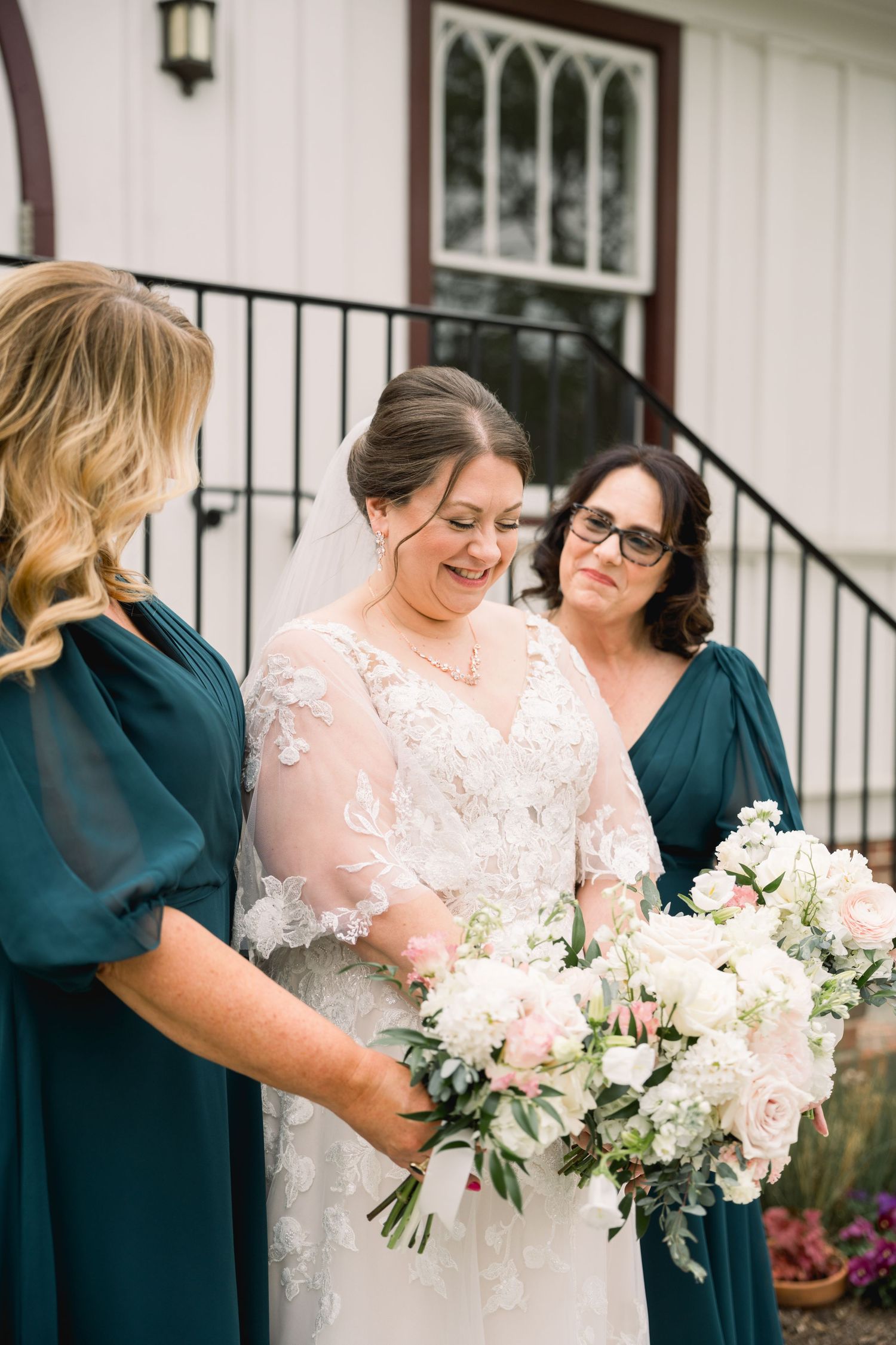 Bride in lace gown laughing with two bridesmaids in teal dresses while holding floral bouquets.