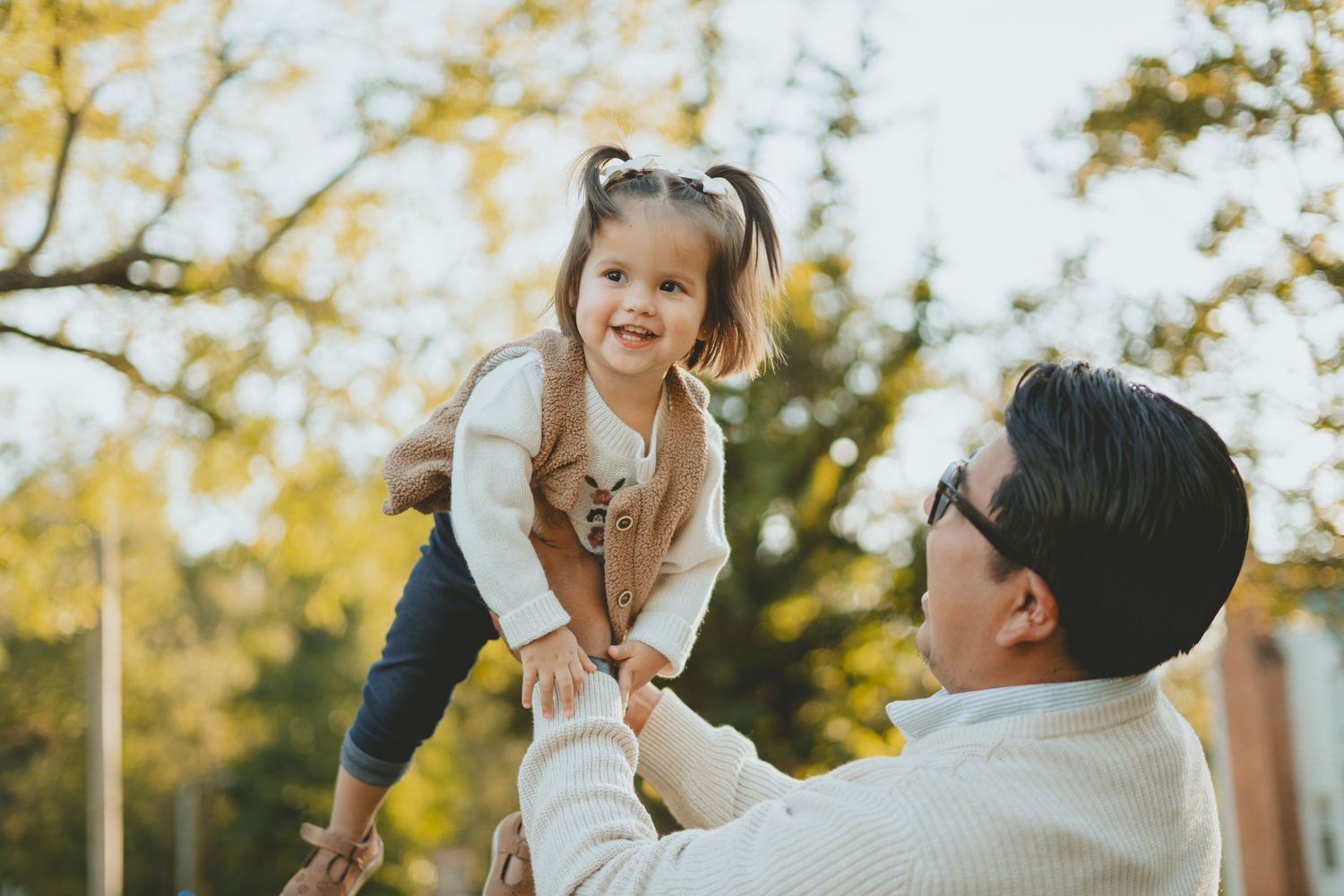 Loving father lifting his smiling toddler daughter into the air outdoors in golden autumn sunlight.