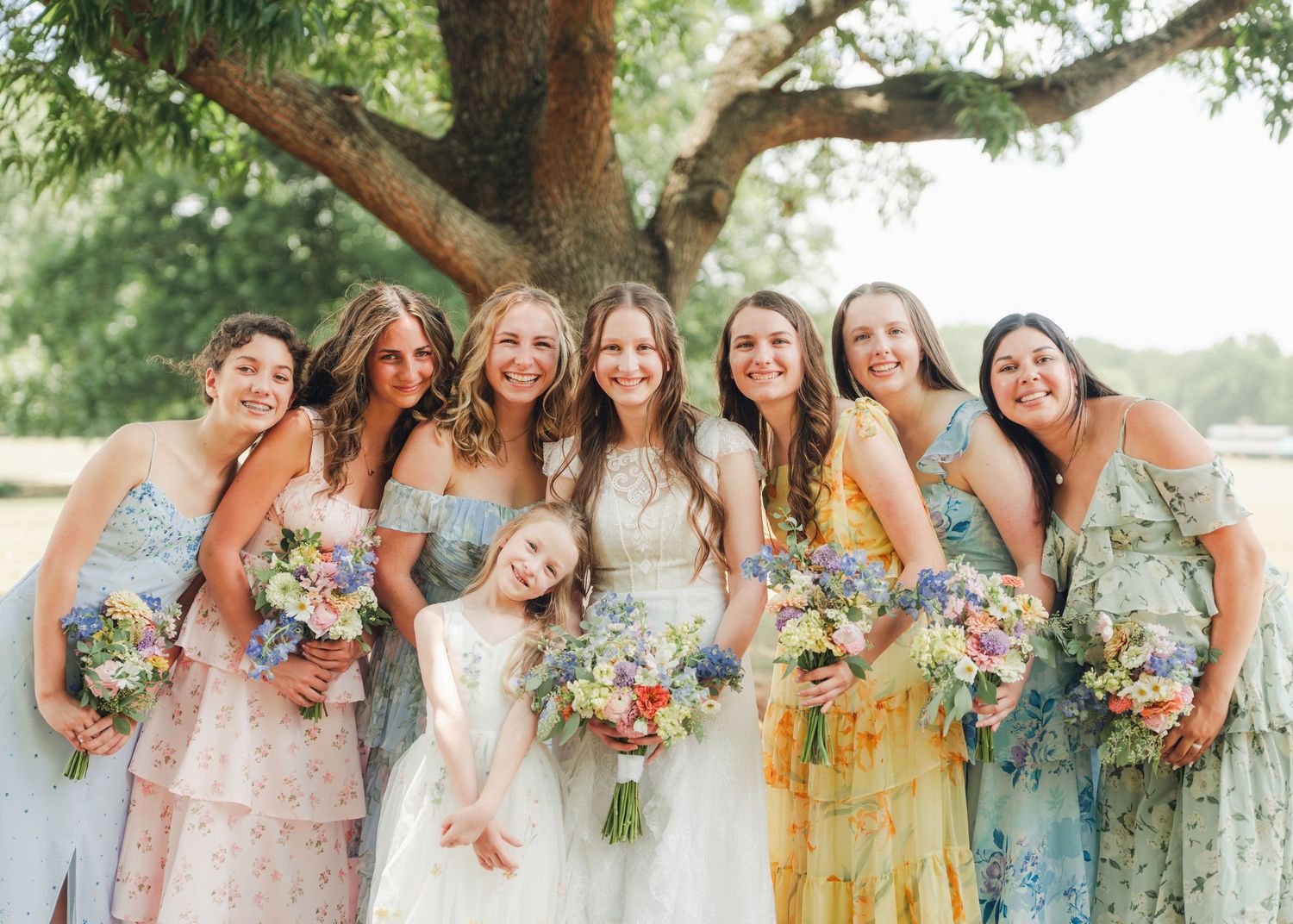 Bride with bridesmaids in colorful floral dresses holding wildflower bouquets under large oak tree.