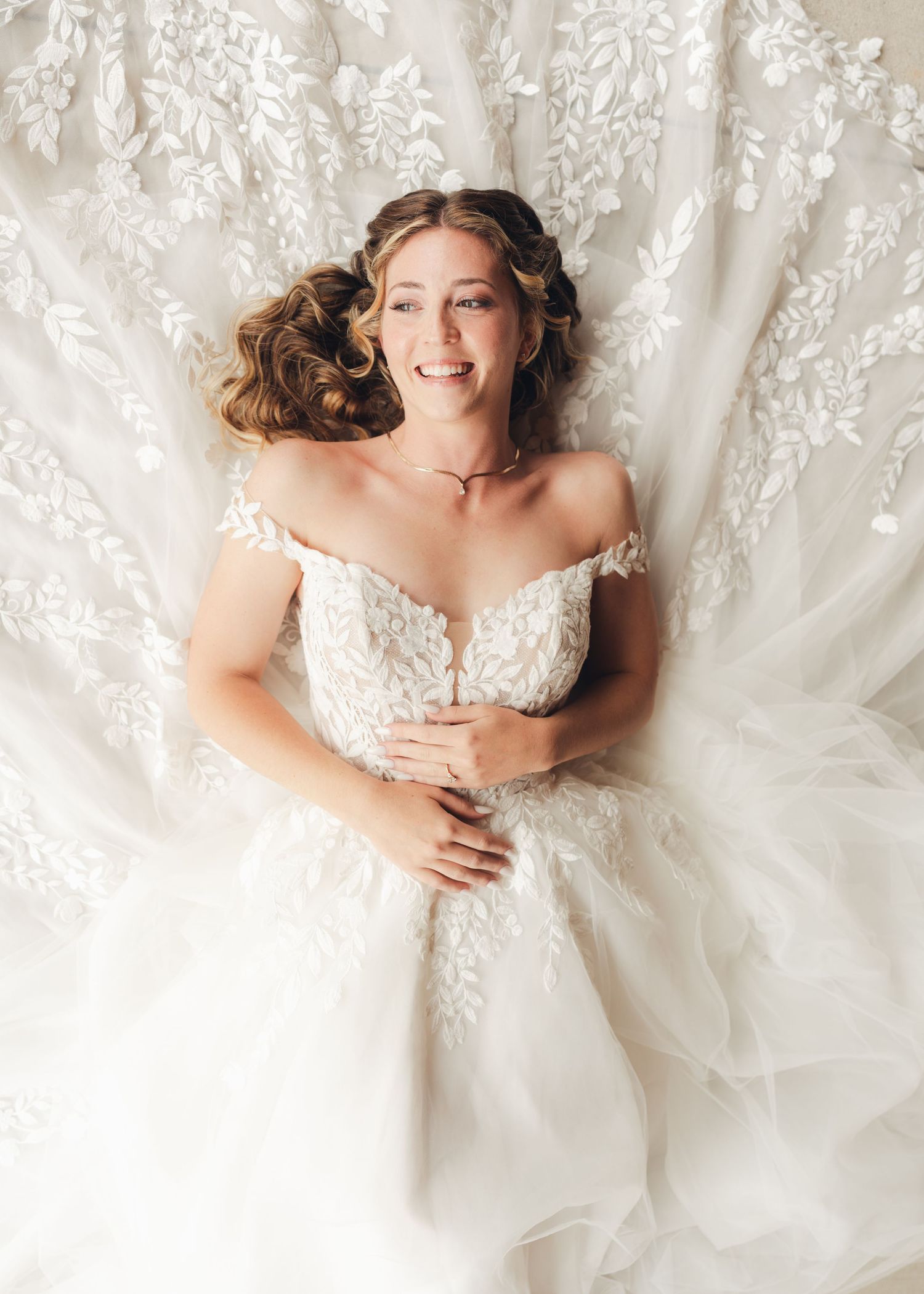 Smiling bride in off-shoulder lace gown lying on her wedding dress skirt from above.