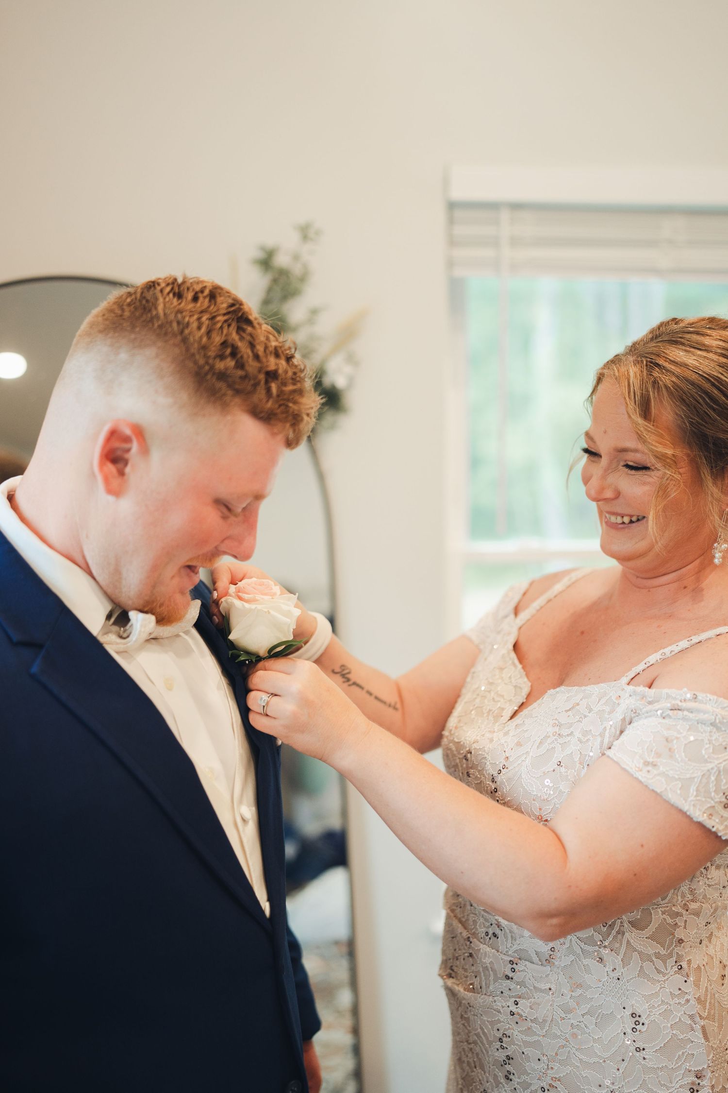 Bride in lace gown pins a white rose boutonniere on groom in navy suit, both smiling during wedding preparations.