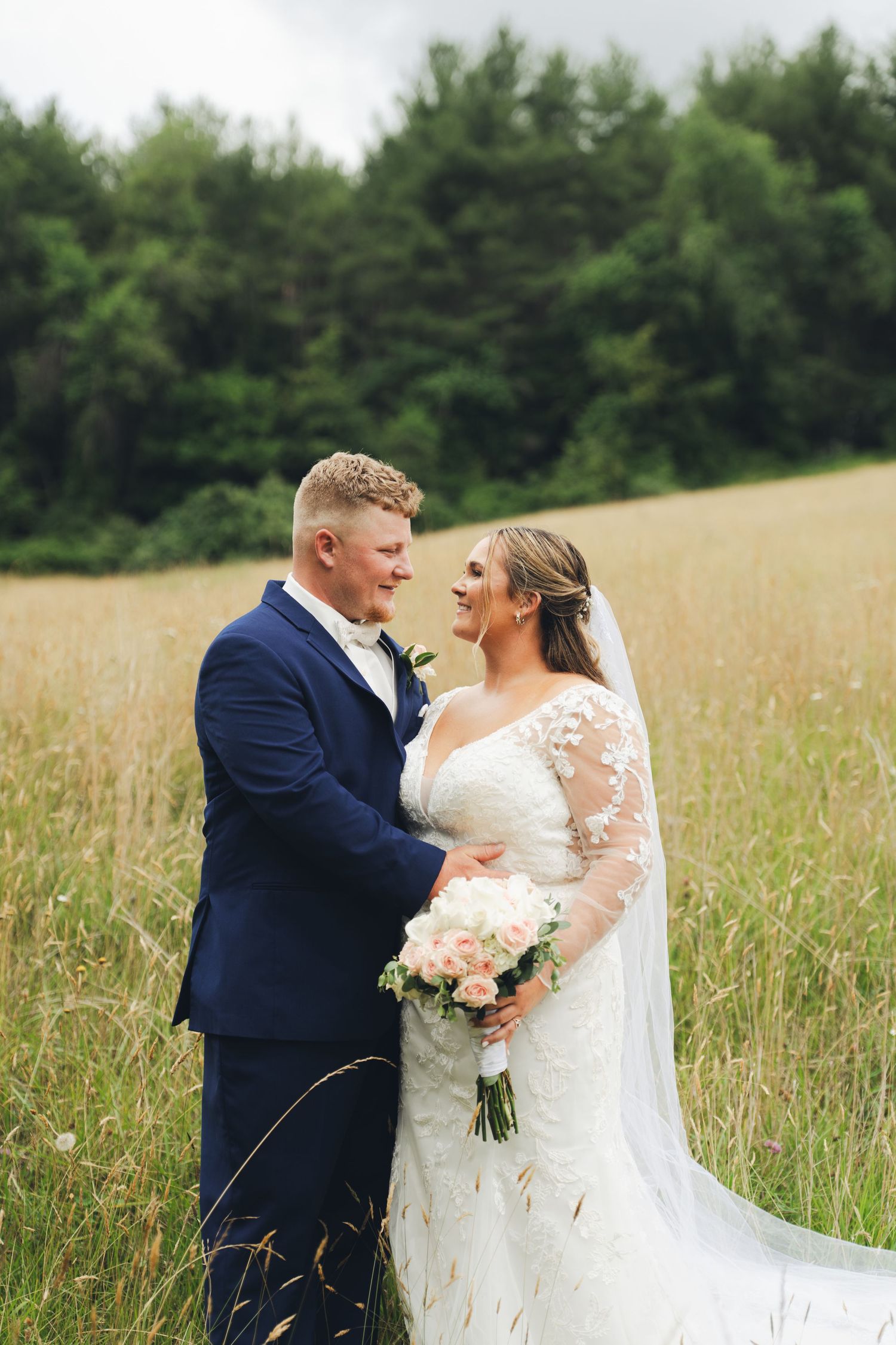 Bride and groom embrace in a golden wheat field, bride holding pink and white bouquet, groom in navy suit.