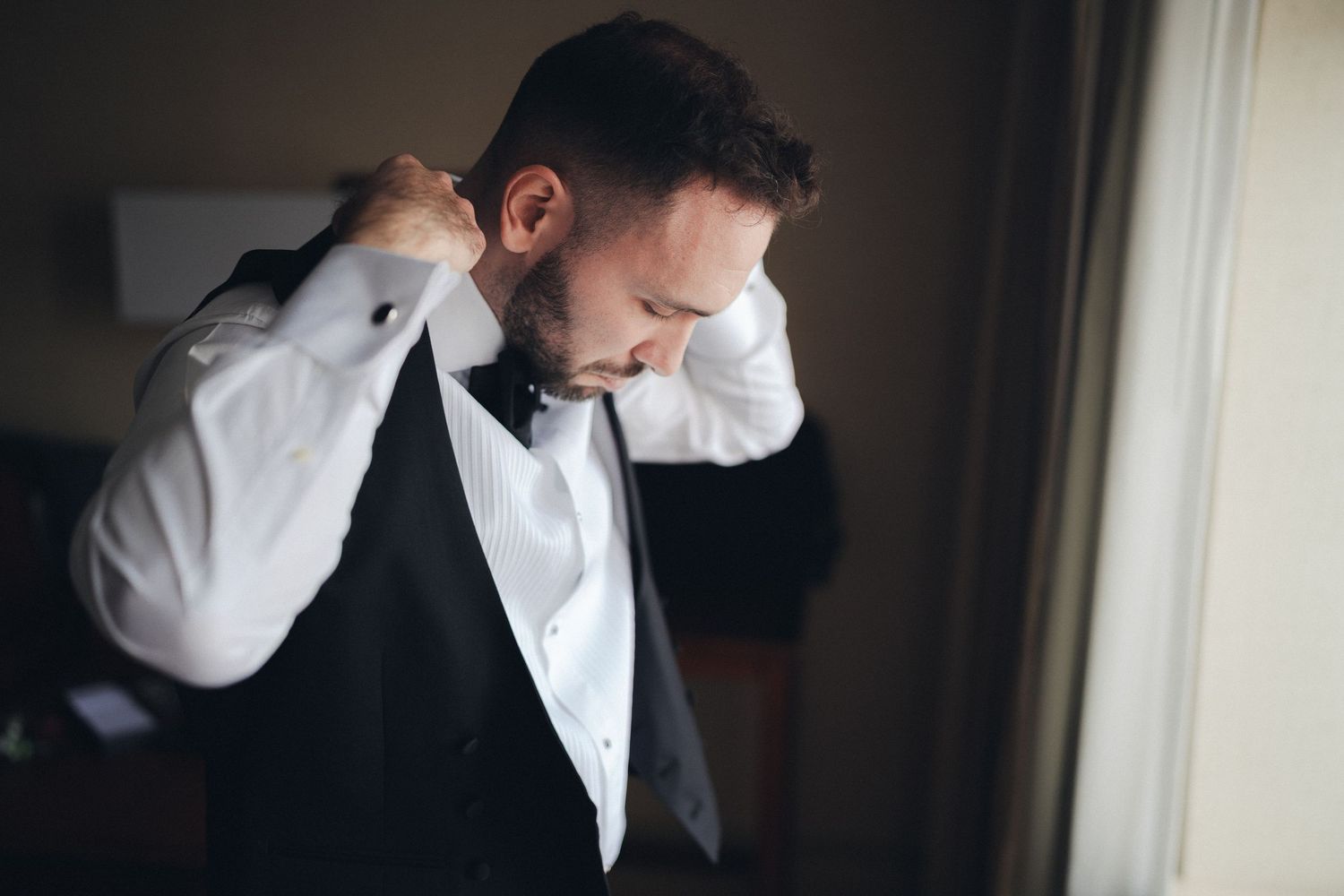 Groom in white shirt and black vest adjusting his bow tie near a window.
