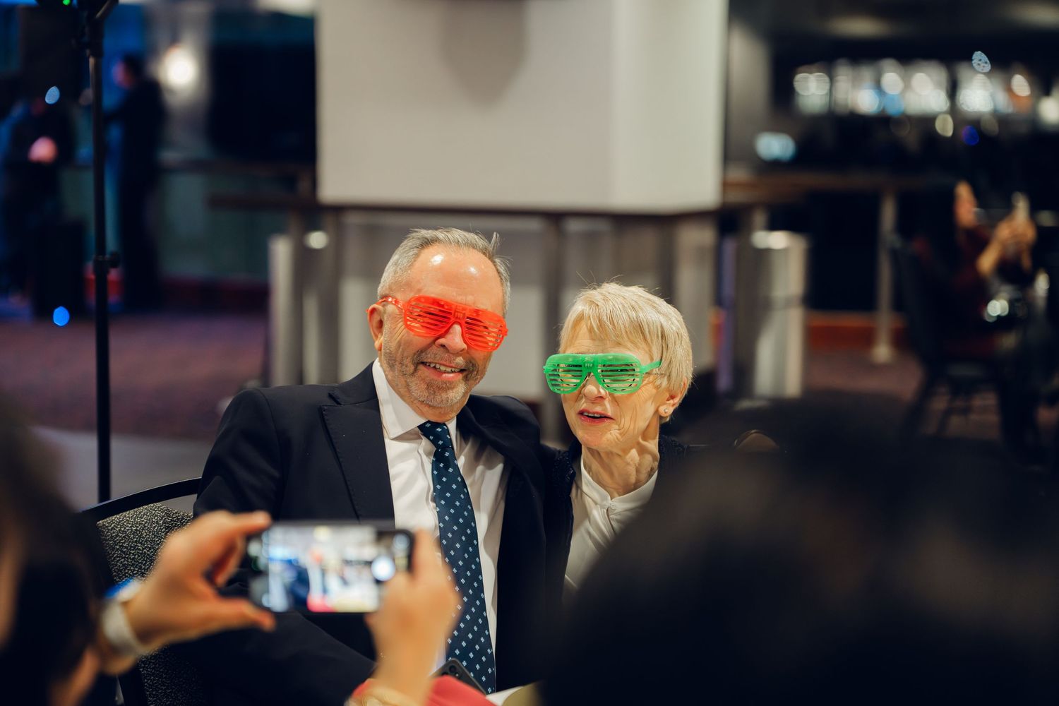 Elderly couple poses playfully wearing oversized colorful novelty glasses at wedding reception.