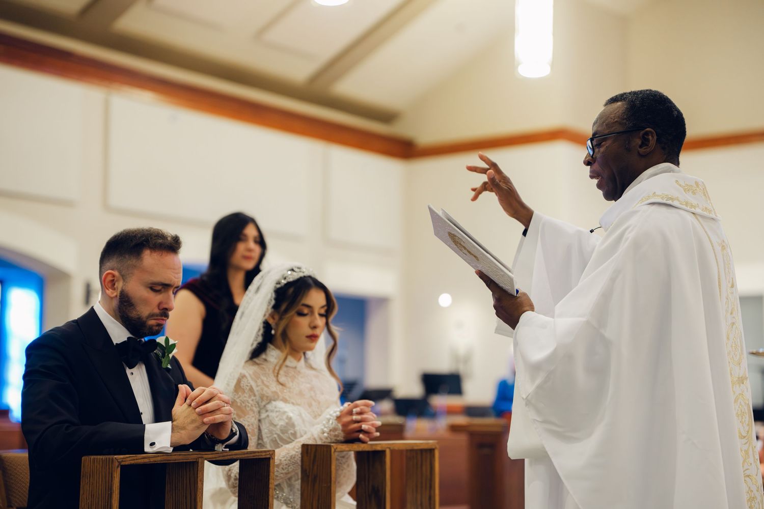 African American priest blesses kneeling bride and groom during Catholic wedding ceremony.