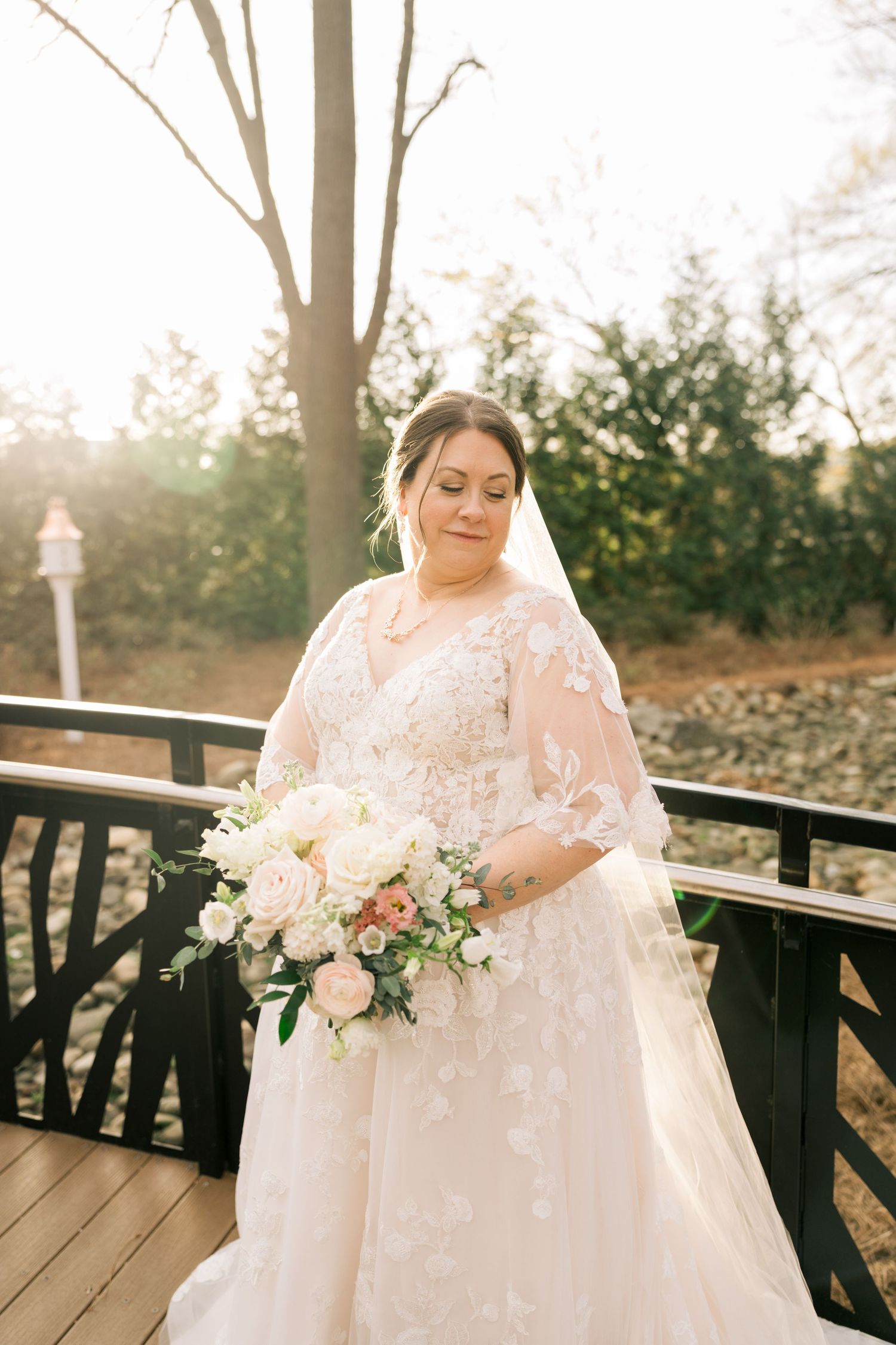 Bride in lace wedding gown holding blush bouquet stands on wooden bridge in golden sunlight.