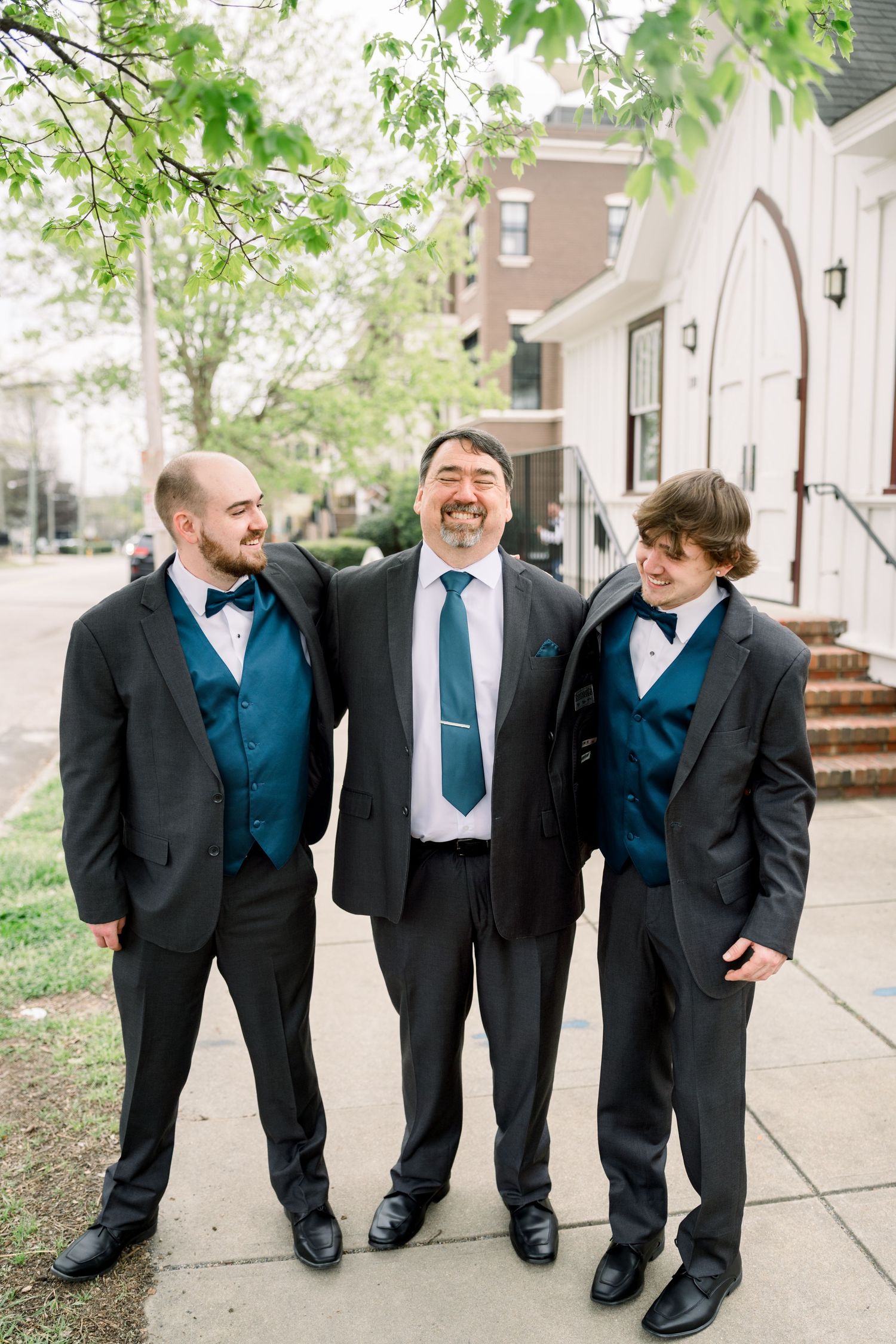 Groom and two groomsmen in black tuxedos with teal vests laughing outside white church.