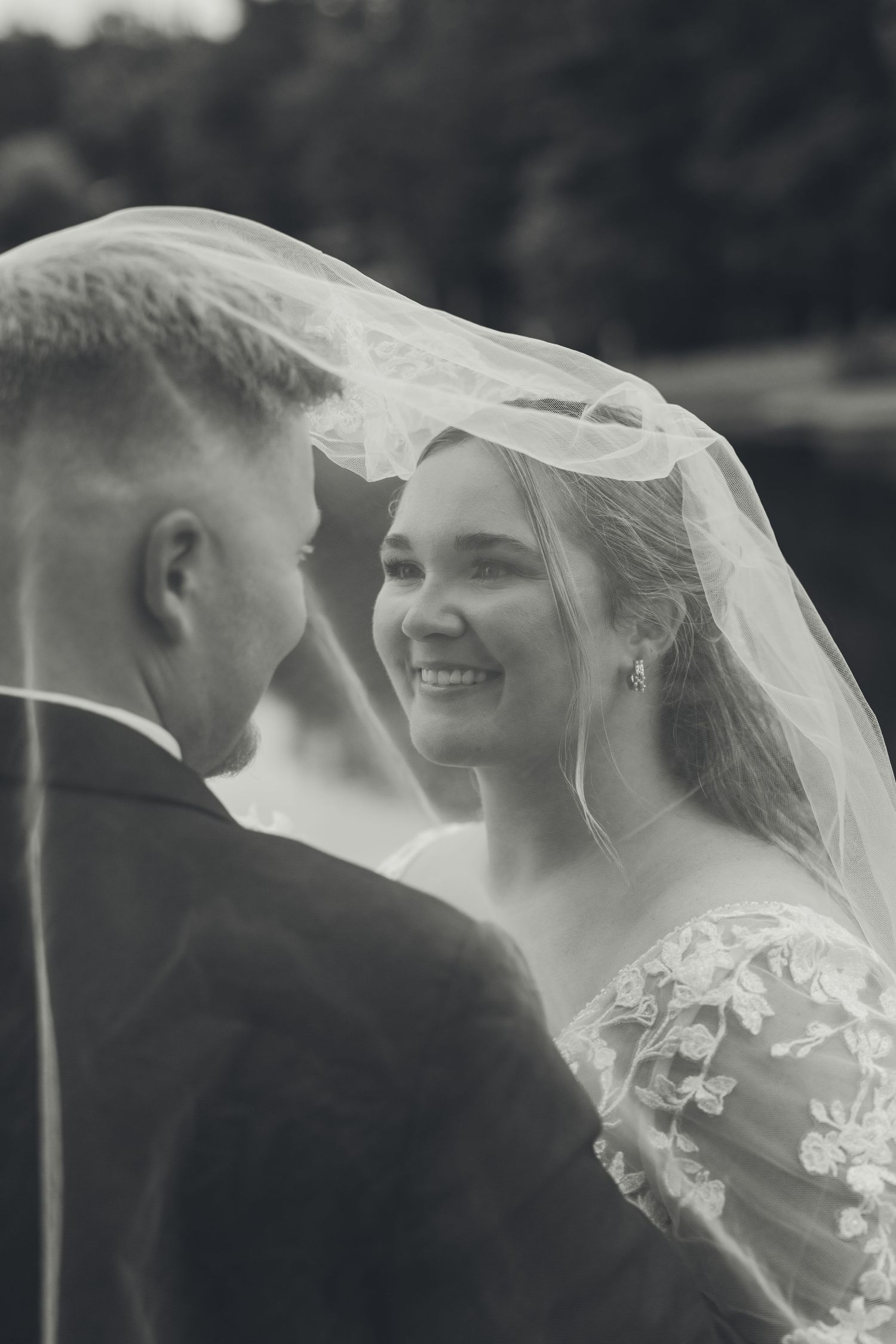 Black and white photo of a smiling bride under veil gazing at groom during romantic wedding ceremony outdoors.