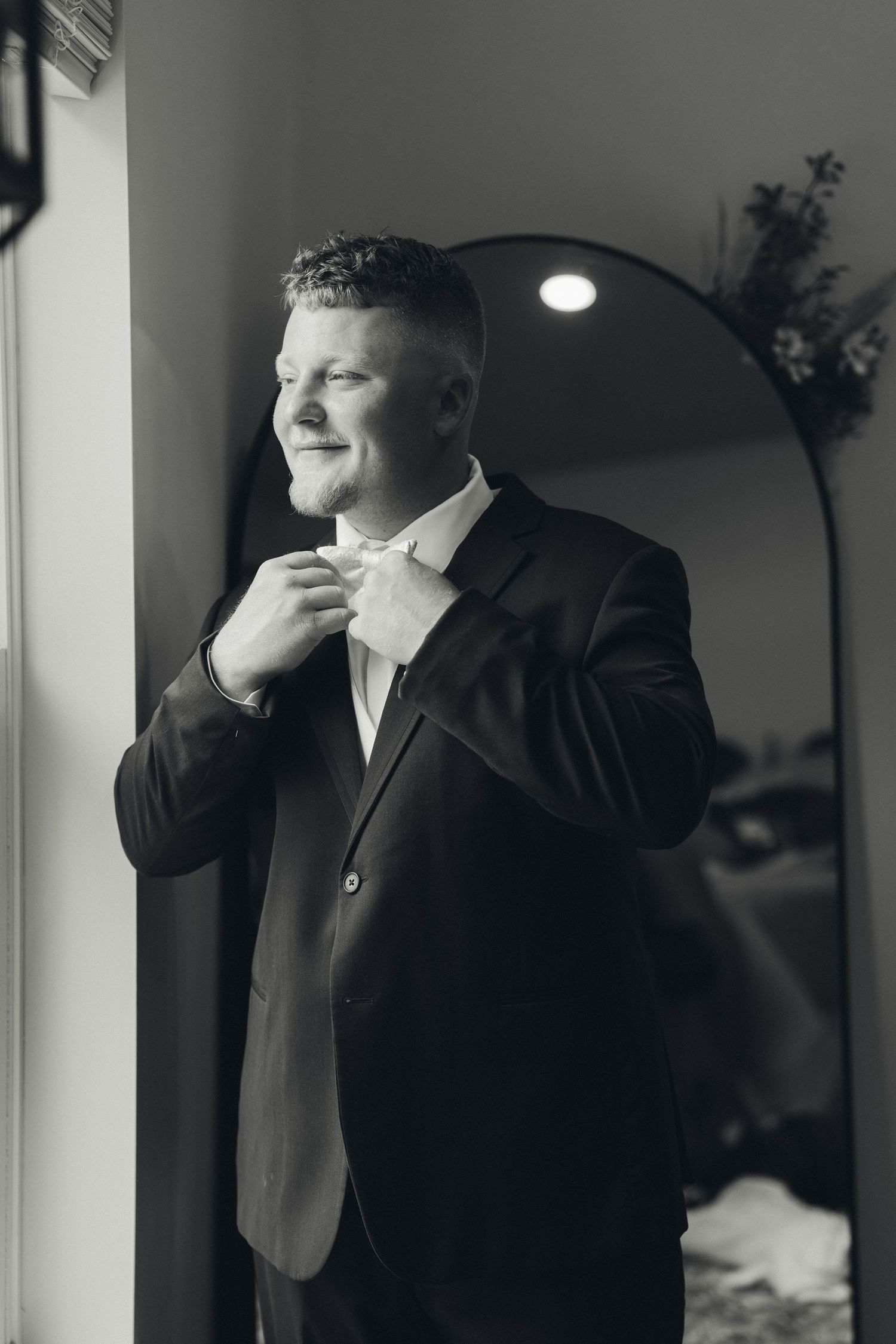 Groom in black suit adjusting bow tie while smiling, standing by window in black and white wedding photo.