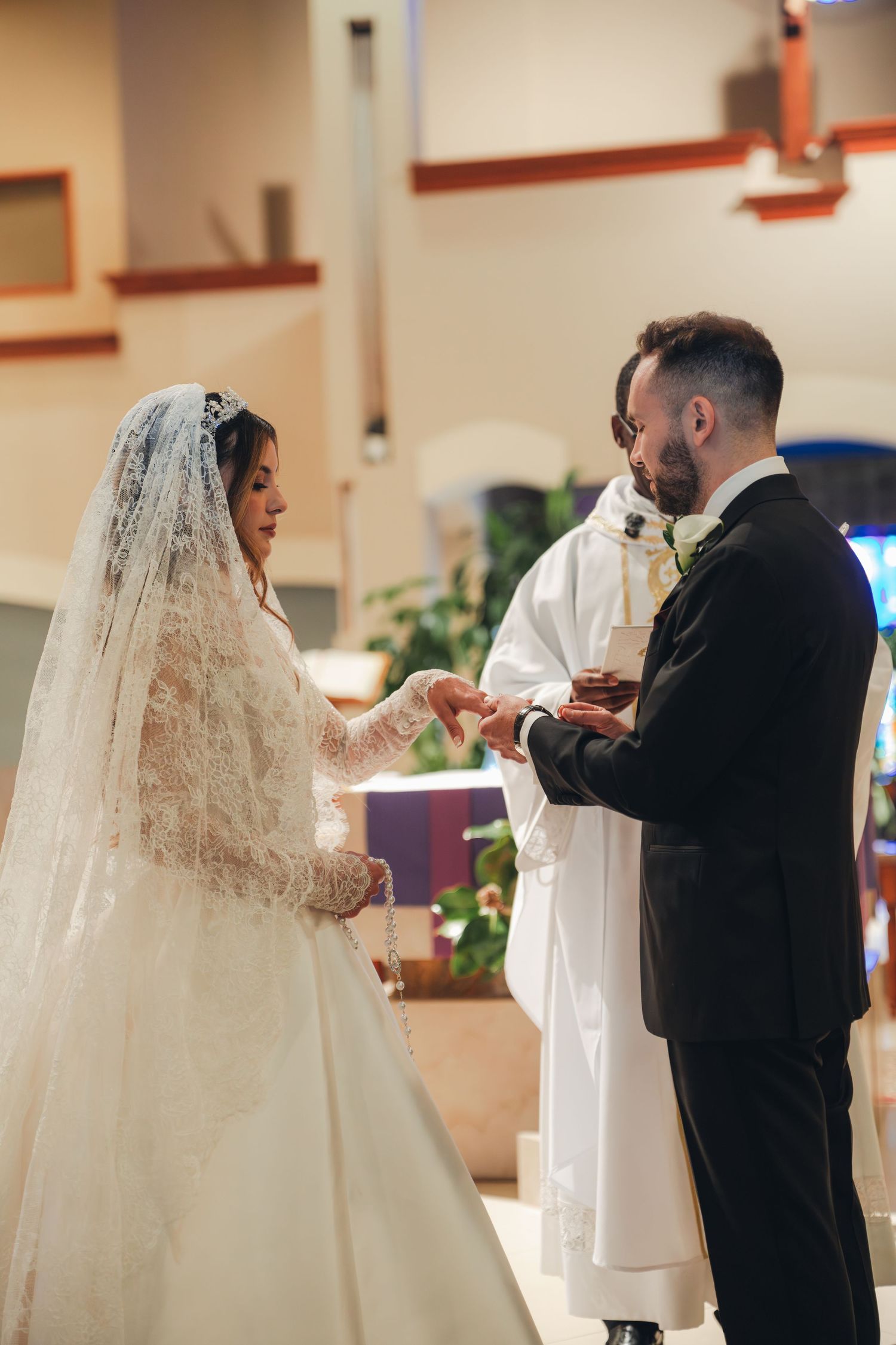 Bride places wedding ring on grooms finger at church altar with priest officiating.