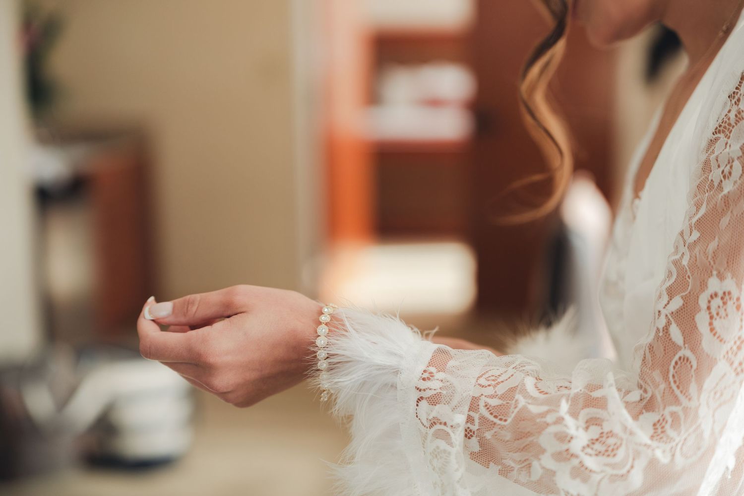 Bride in white lace robe fastening a pearl bracelet on her wrist during wedding preparations.