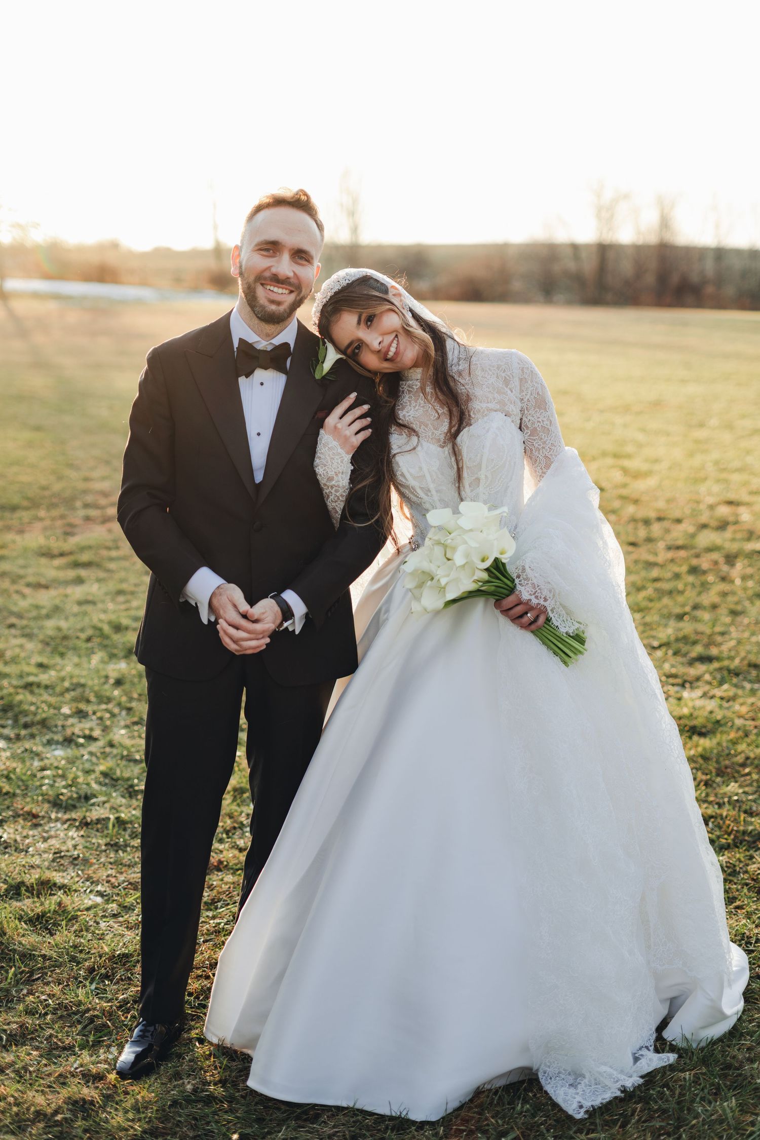 Smiling bride and groom pose together at golden hour in a sunlit field, bride holding white calla lily bouquet.