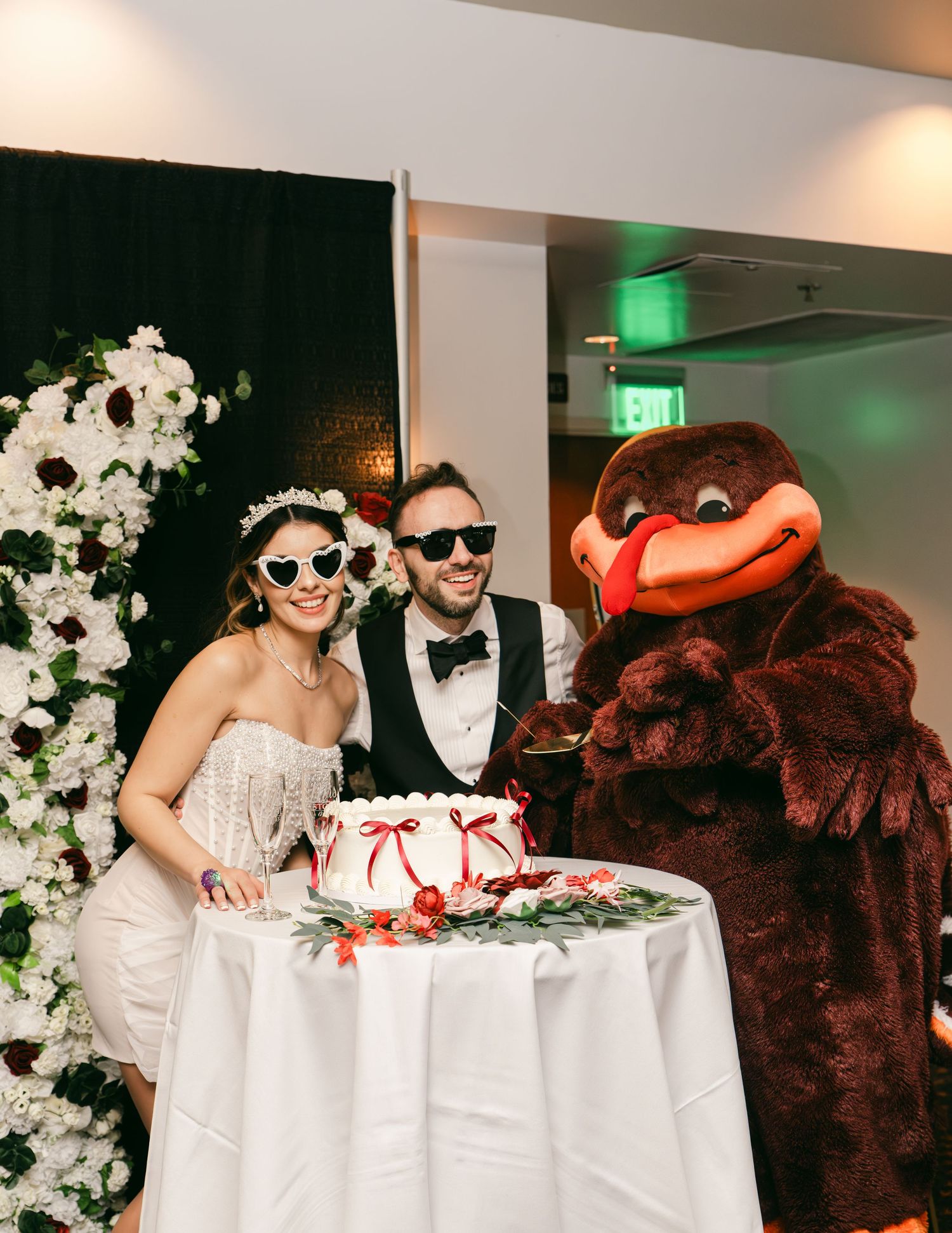 Bride and groom wearing heart sunglasses pose with a mascot costume beside white wedding cake at fun reception.