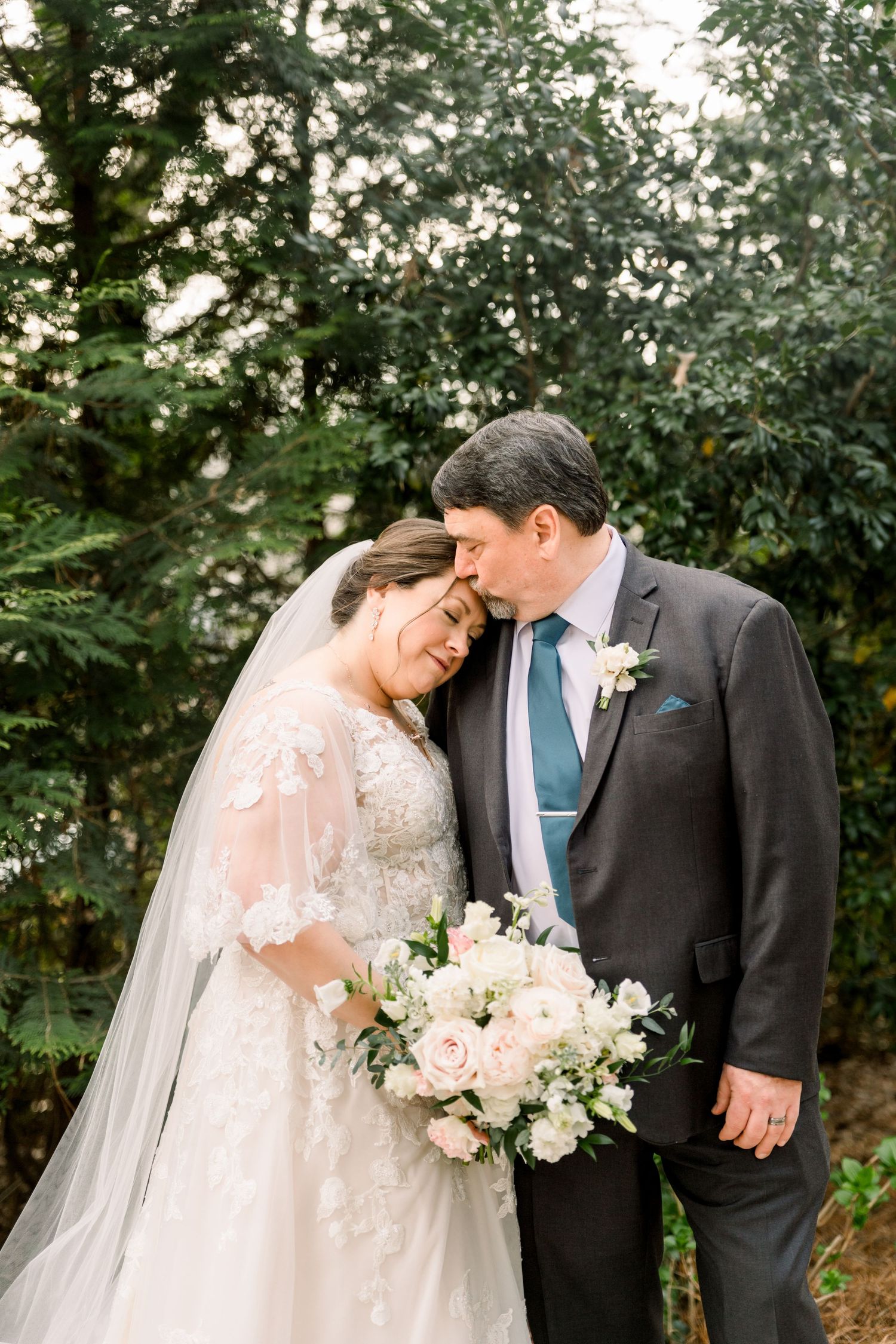 Bride and groom touch foreheads tenderly while holding white floral bouquet outdoors among green trees.