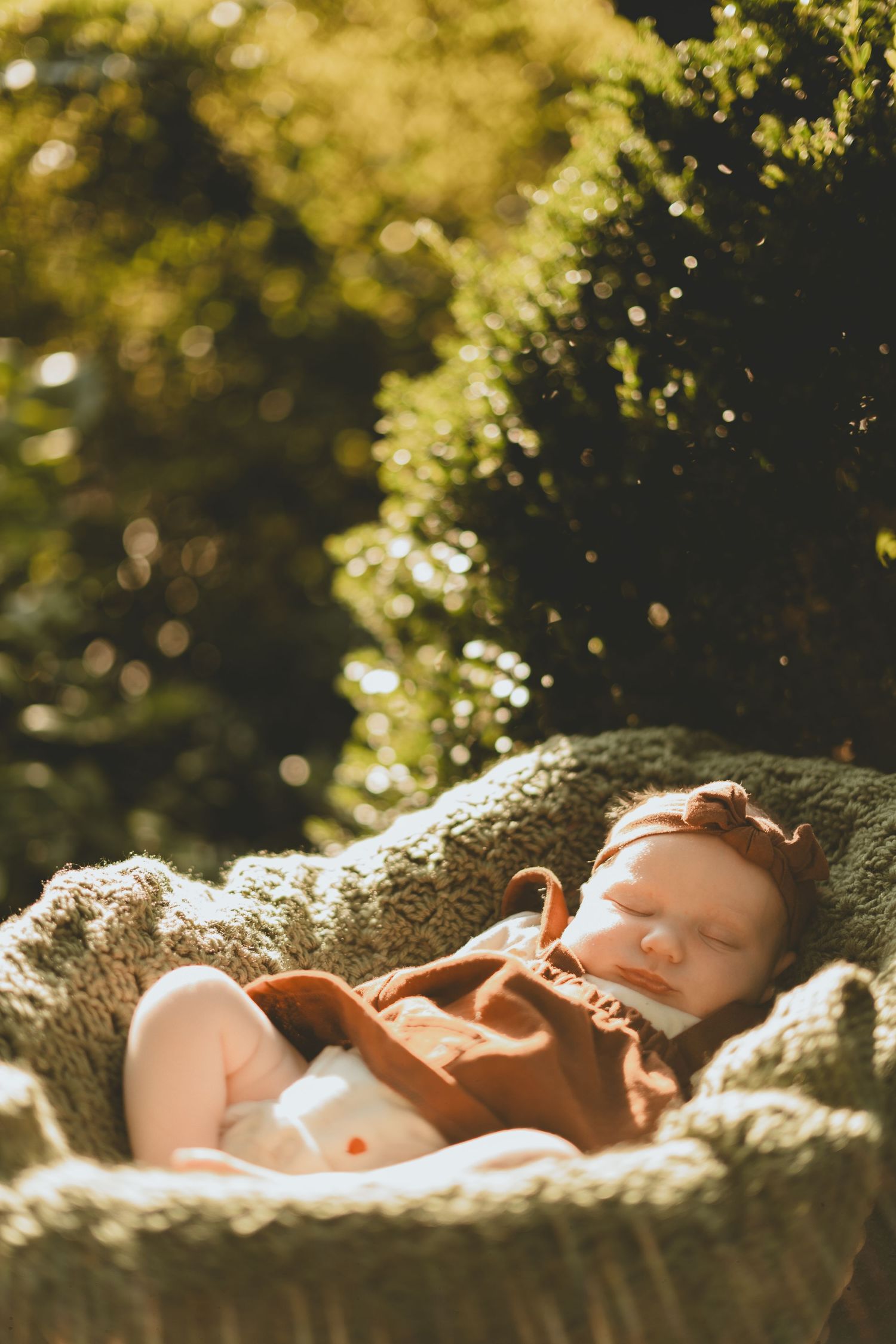A sleeping newborn baby in brown overalls rests peacefully in a knit basket surrounded by greenery.
