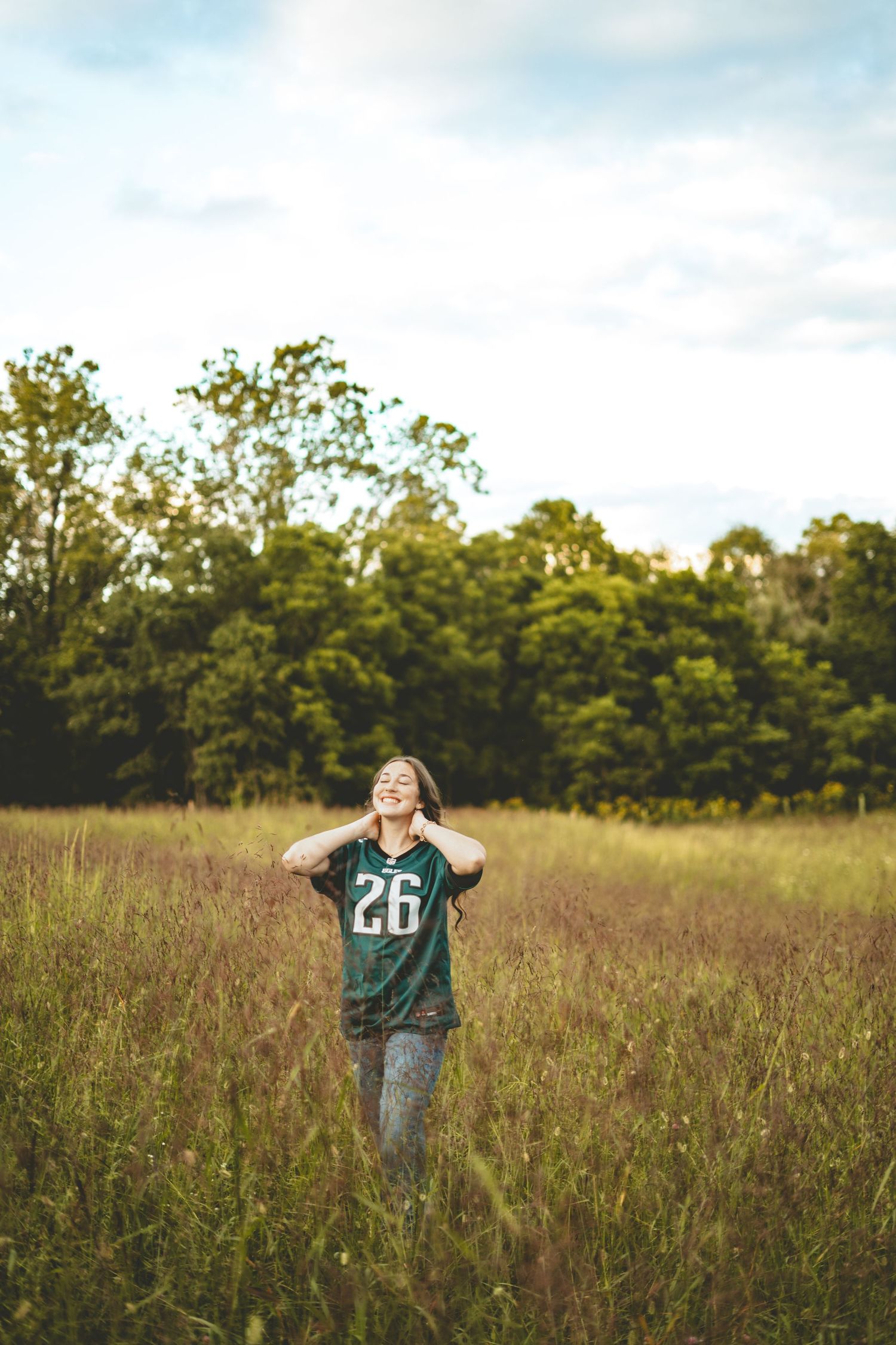 Teen girl in green Eagles jersey number 26 stands in tall grass field with eyes closed.