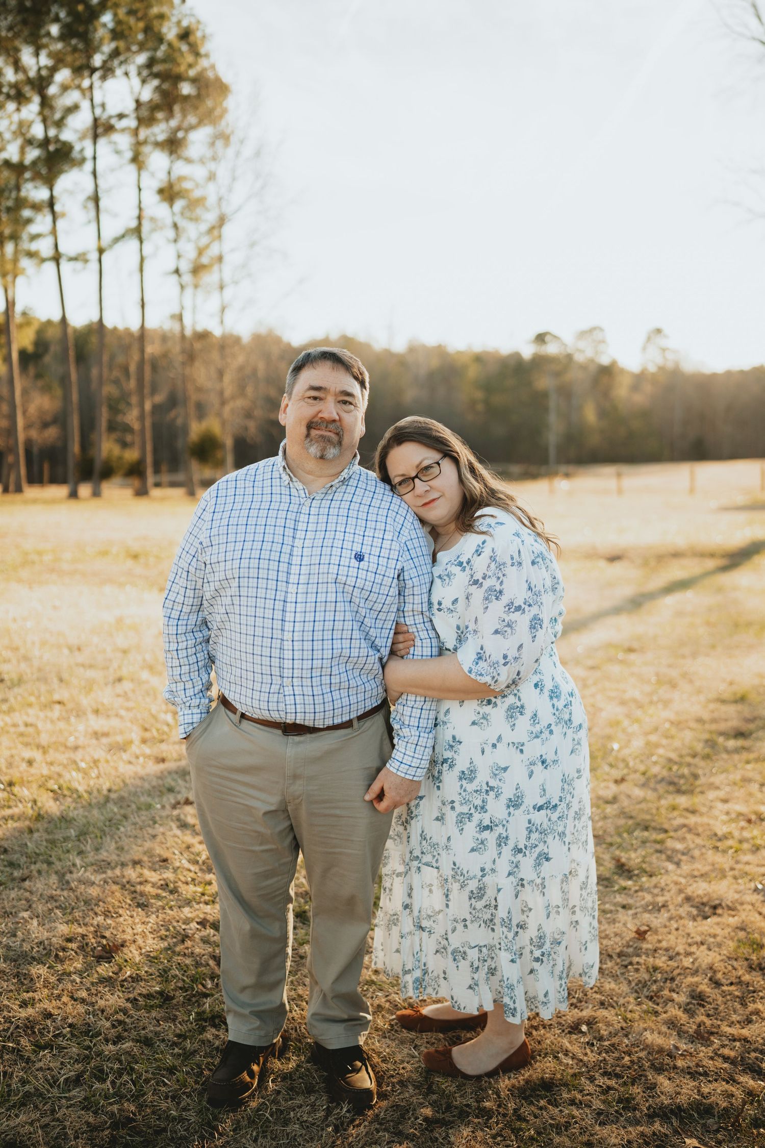 Couple posing together in a sunny field, woman in floral dress leaning on man in blue plaid shirt.
