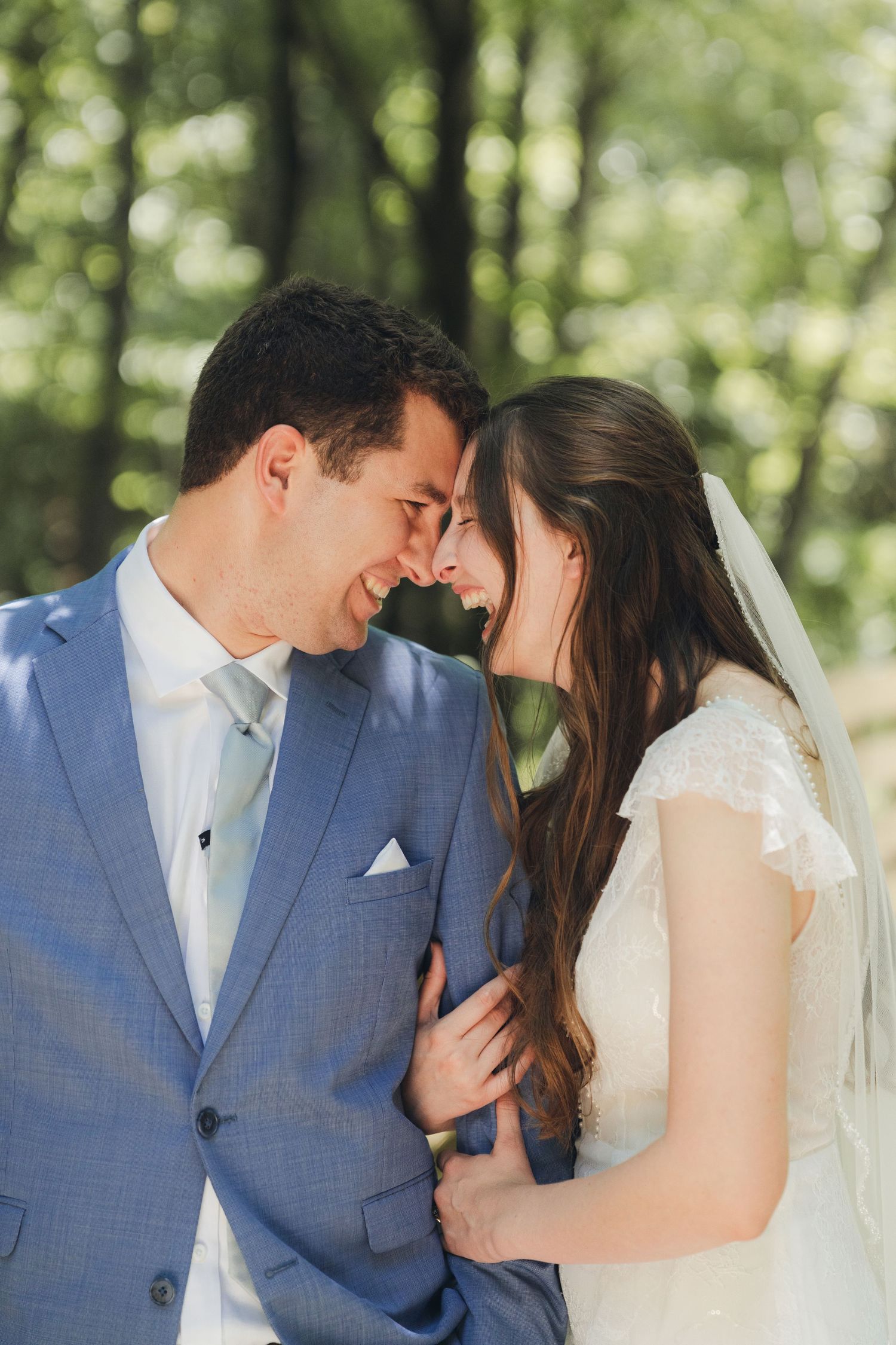 Smiling bride and groom touching foreheads tenderly in sun-dappled green outdoor setting.