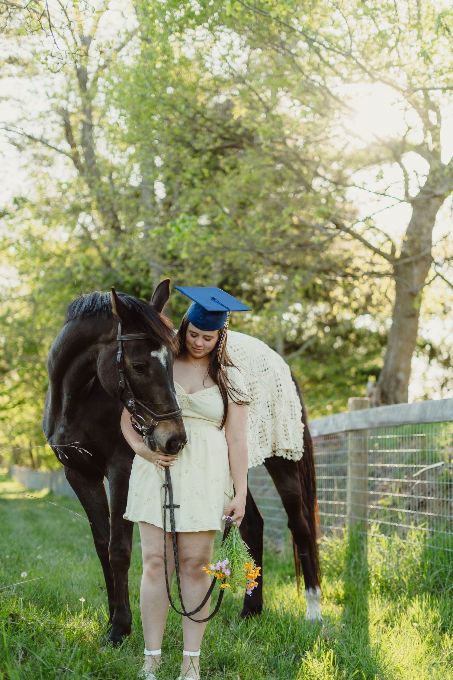 Graduate in blue cap with dark horse in sunny green field, holding wildflowers and bridle.