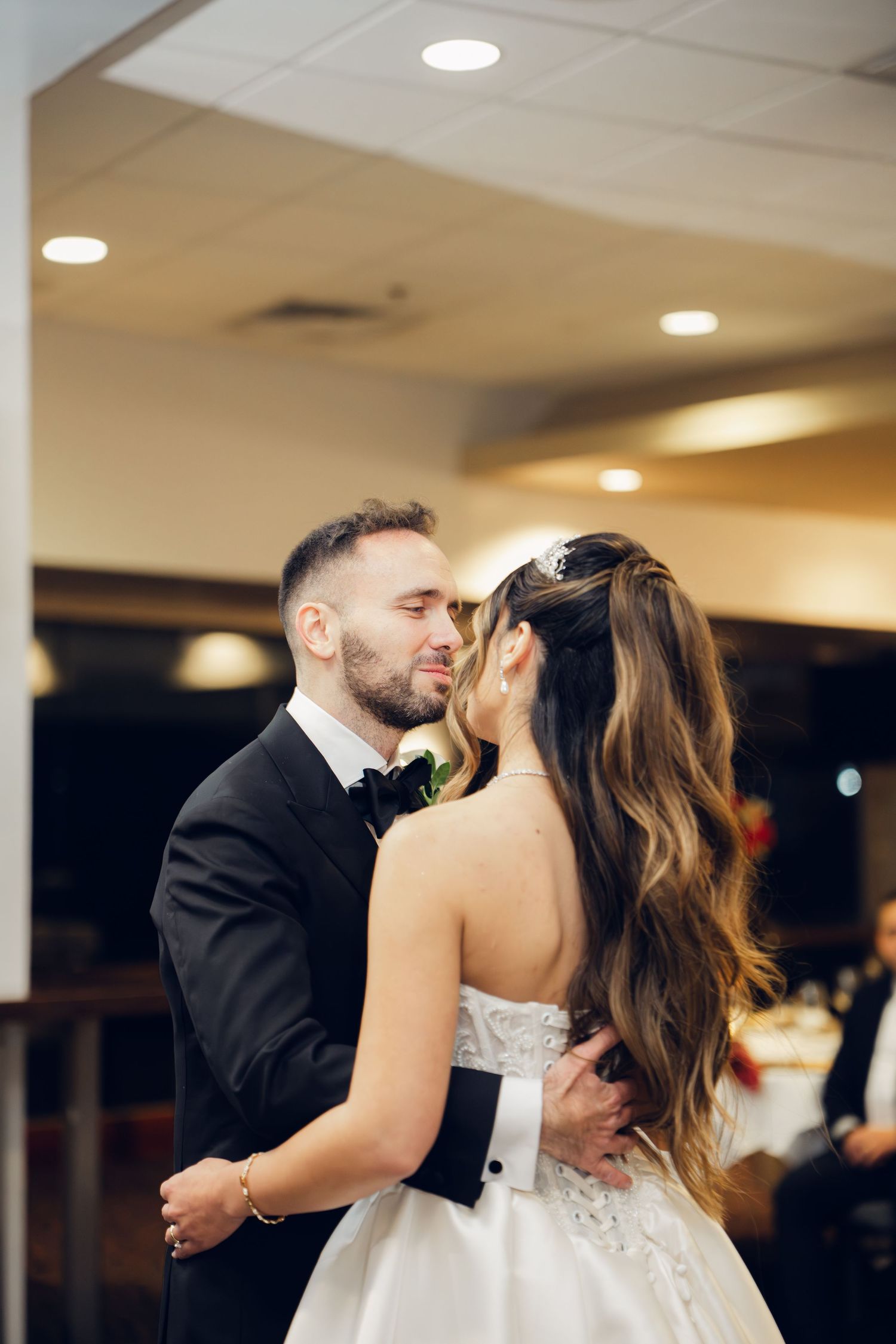 Groom tenderly embraces bride during first dance at indoor wedding reception.