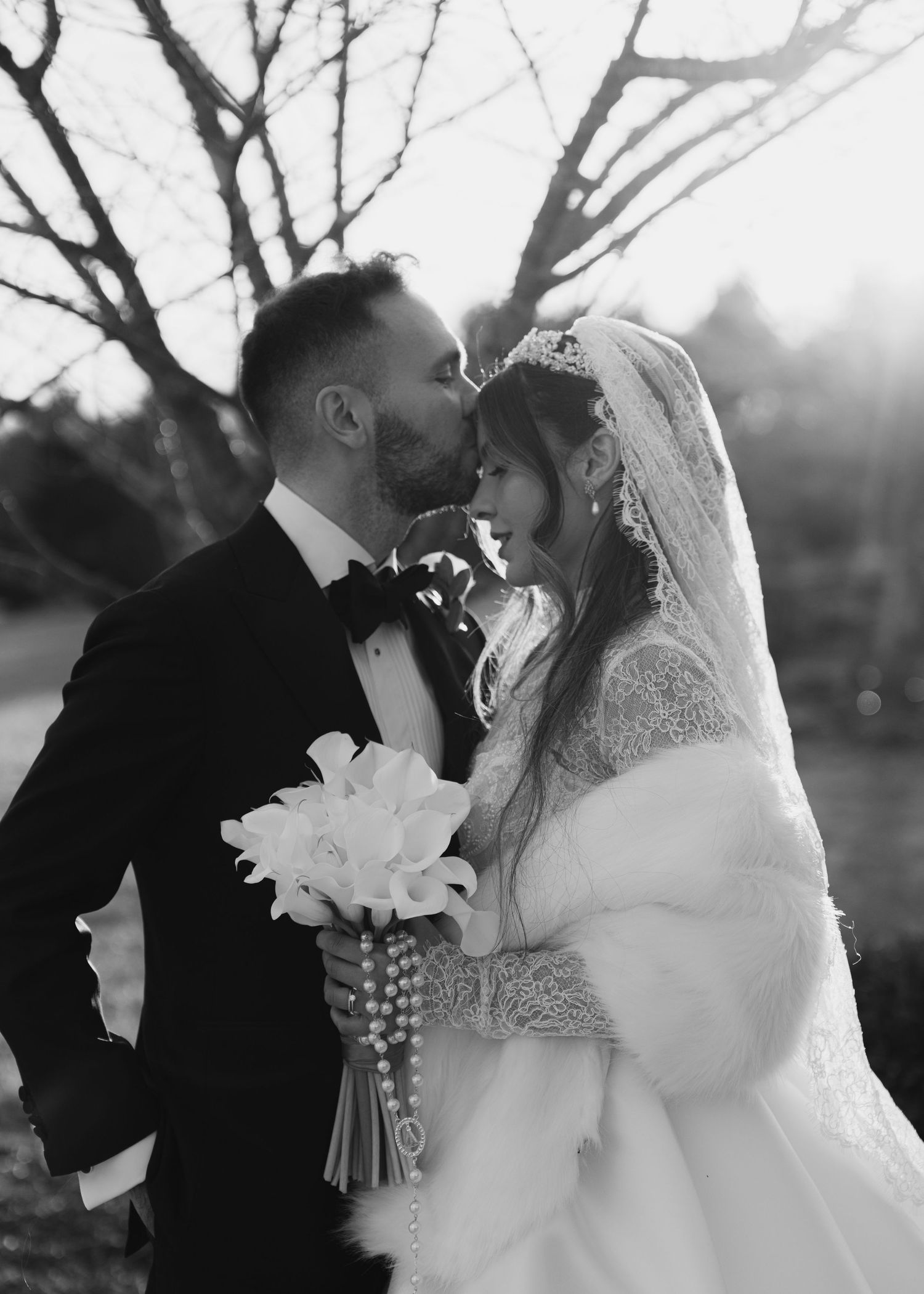 Black and white romantic photo of bride and groom kissing outdoors, bride holding calla lilies and rosary beads.