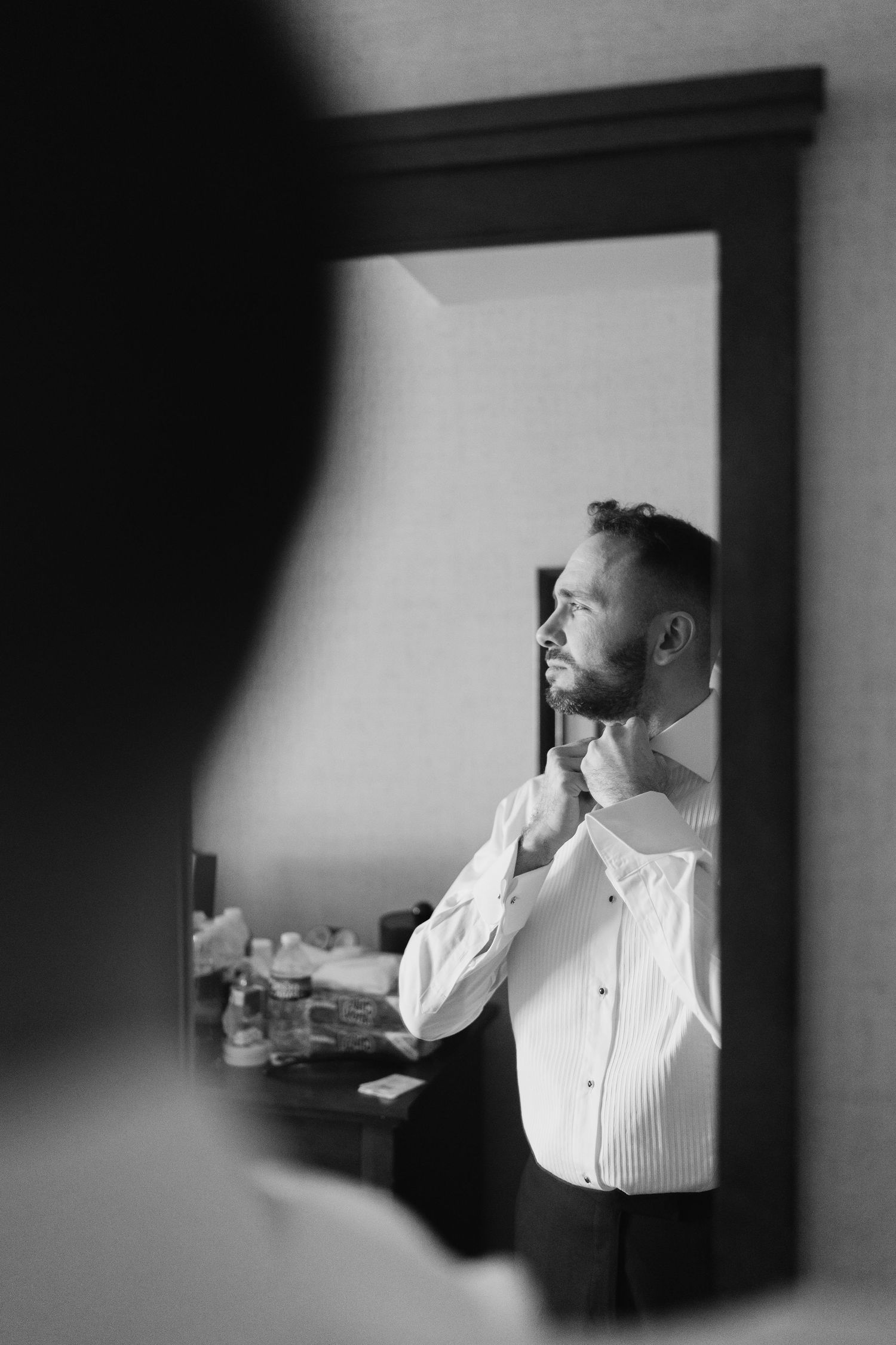 Black and white reflection of groom adjusting collar in mirror while dressing.