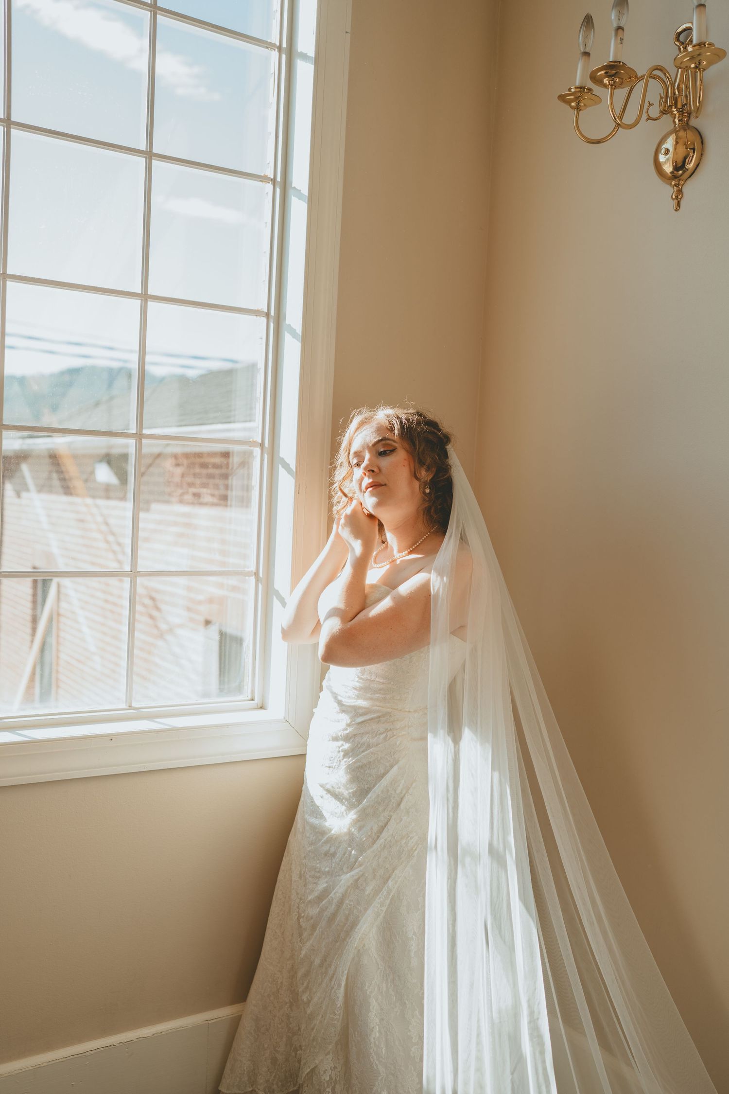 Radiant bride in lace wedding gown and cathedral veil stands by sunlit window with gold wall sconce nearby.