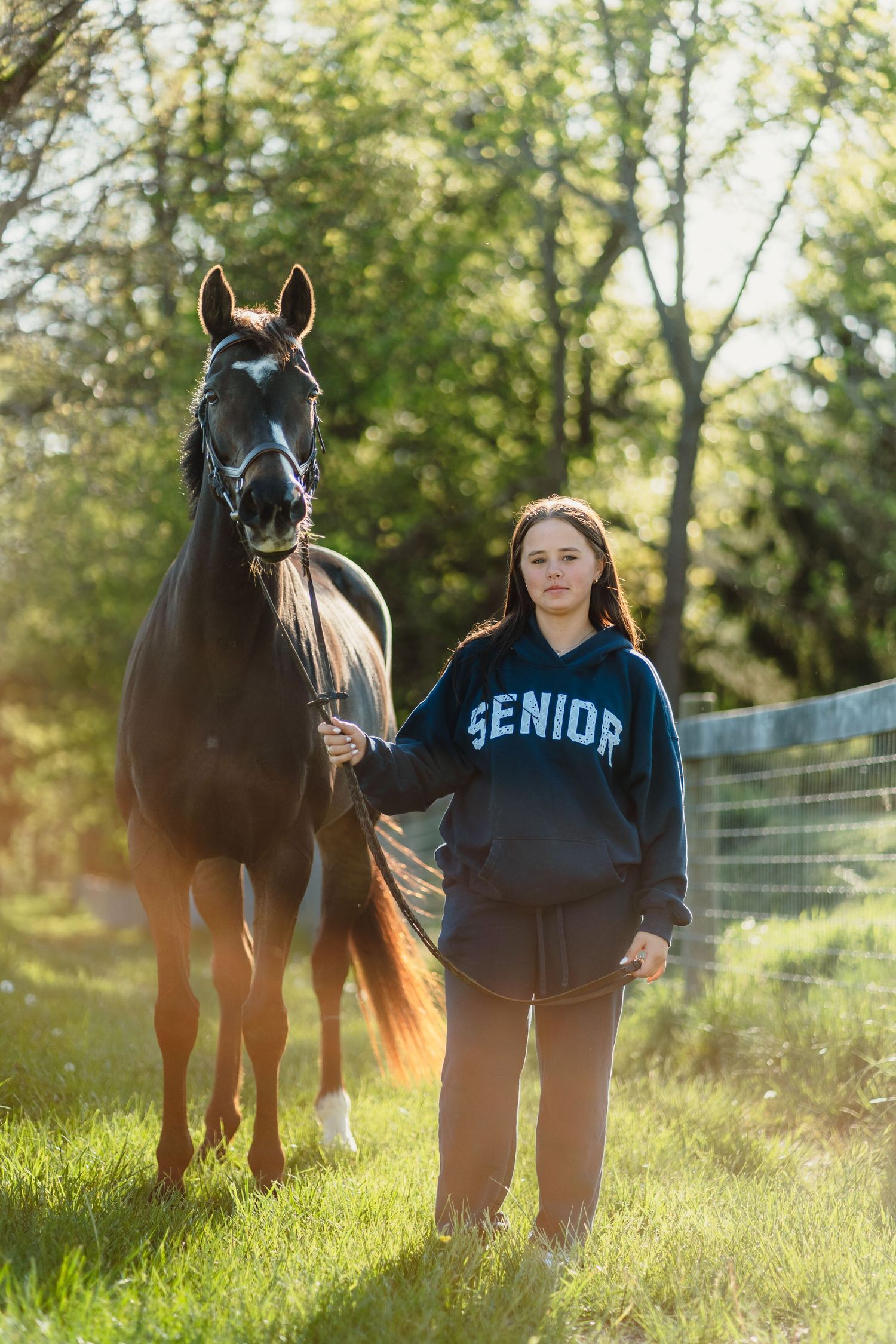 A teenage girl in a Senior hoodie stands beside a dark horse in a sunlit green pasture.