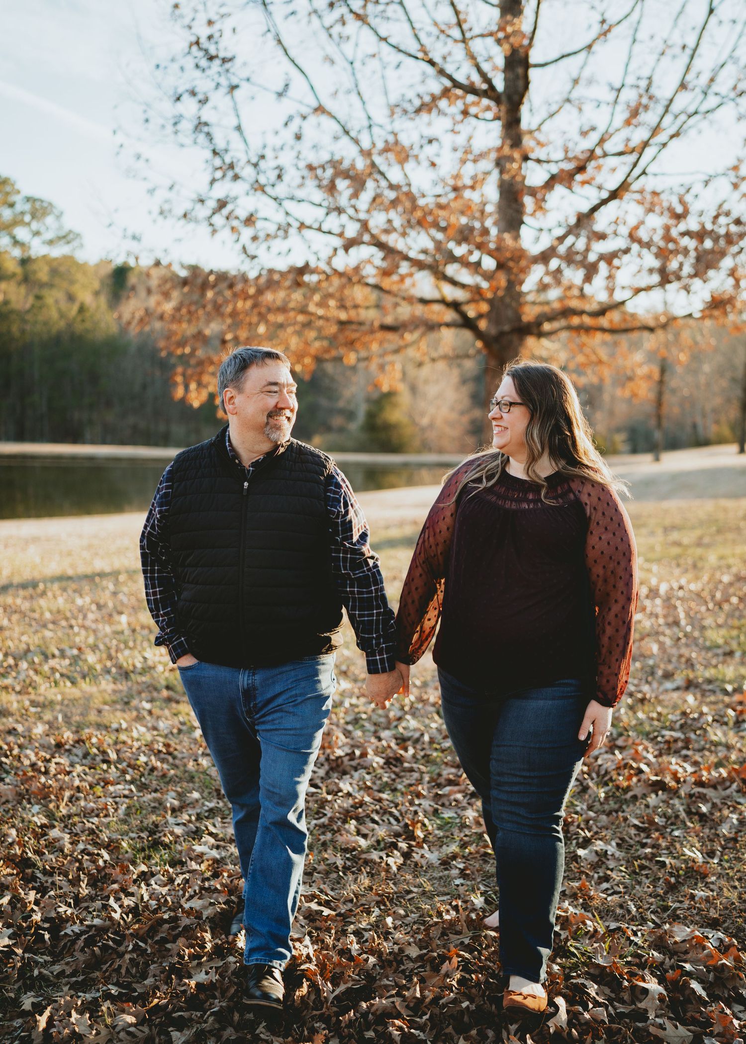 Couple holding hands and walking through autumn leaves near a pond.