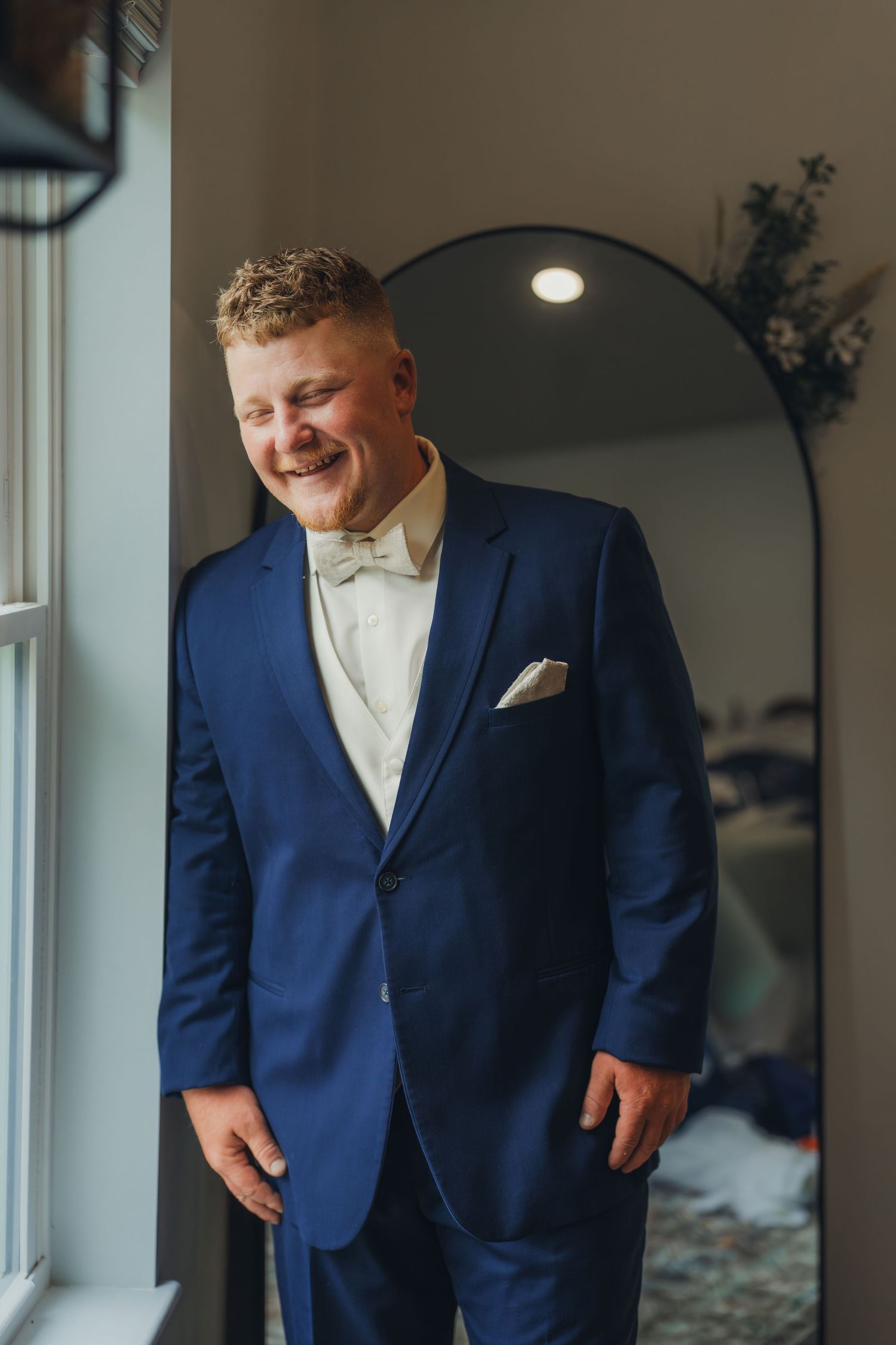 Smiling groom in navy blue suit with white bow tie and pocket square standing by window on wedding day.