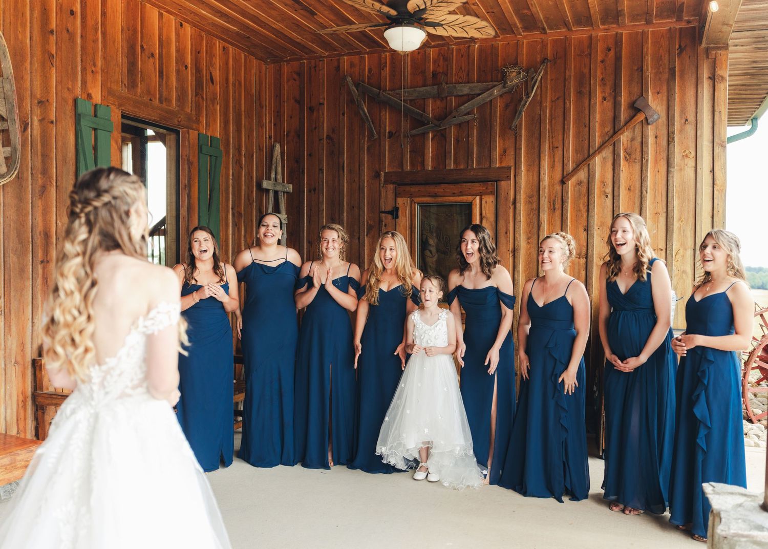Bridesmaids in navy blue gowns react with joy as bride reveals her wedding dress in a rustic barn venue.