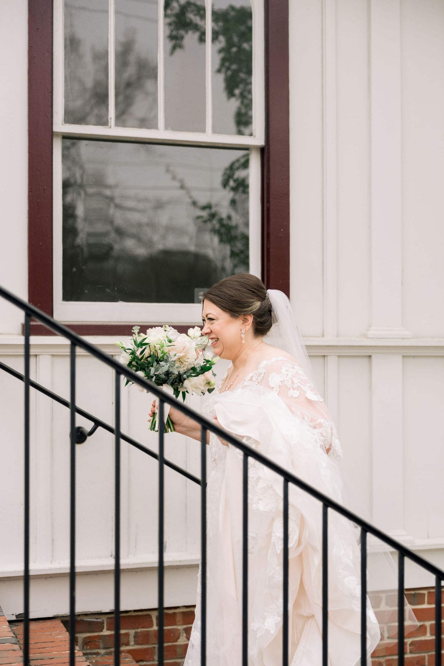 Joyful bride in lace gown and veil laughs while holding white bouquet on outdoor staircase.