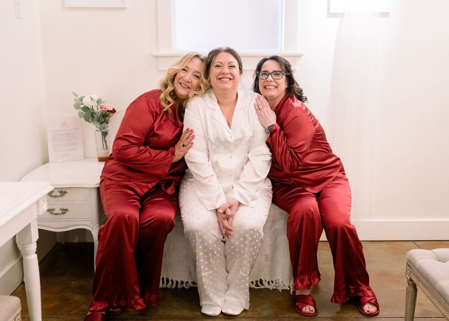 Bride in white robe sitting between two bridesmaids in red satin pajamas smiling.