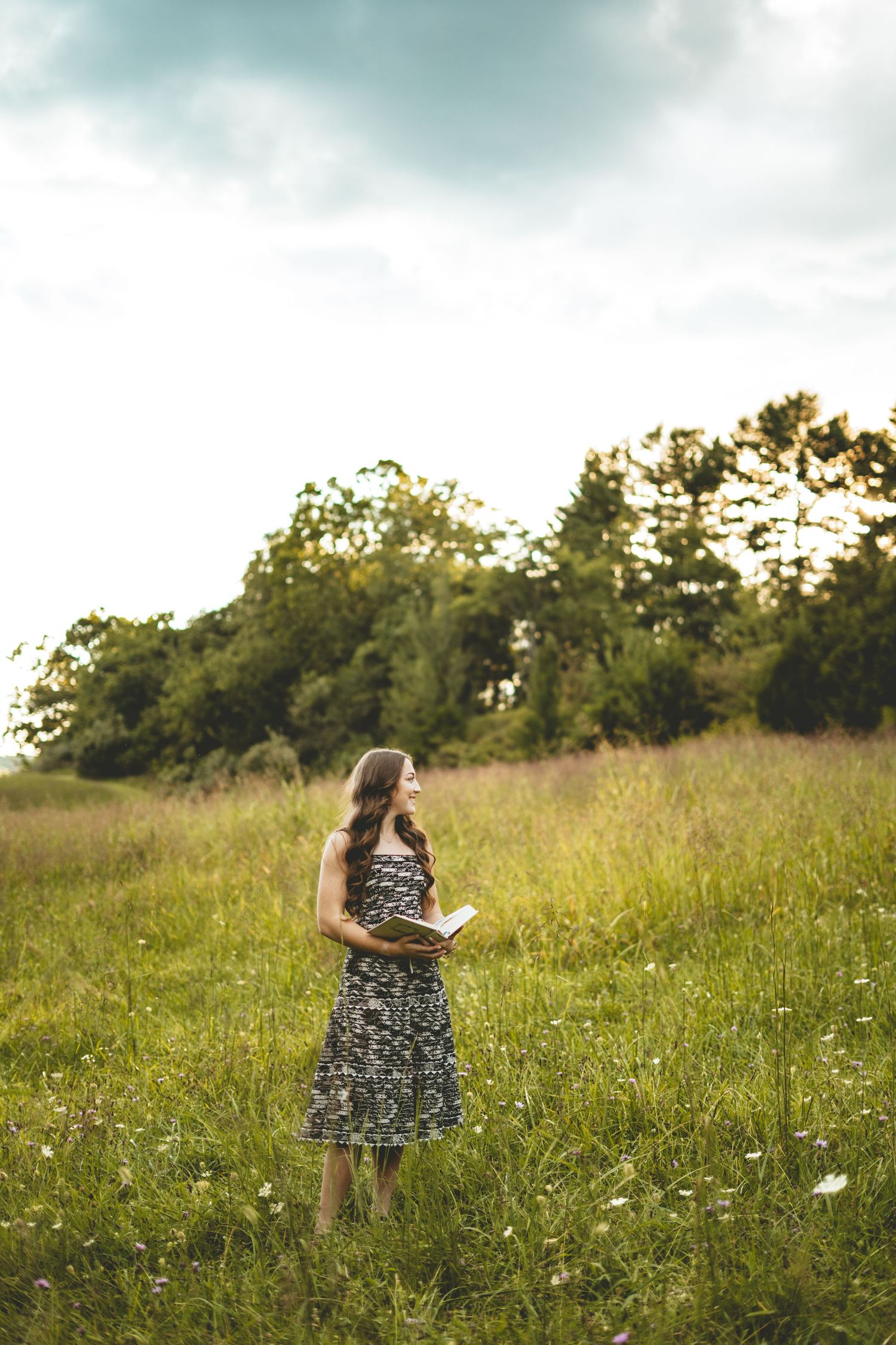 Young woman in floral dress stands in open meadow holding book under dramatic cloudy sky.