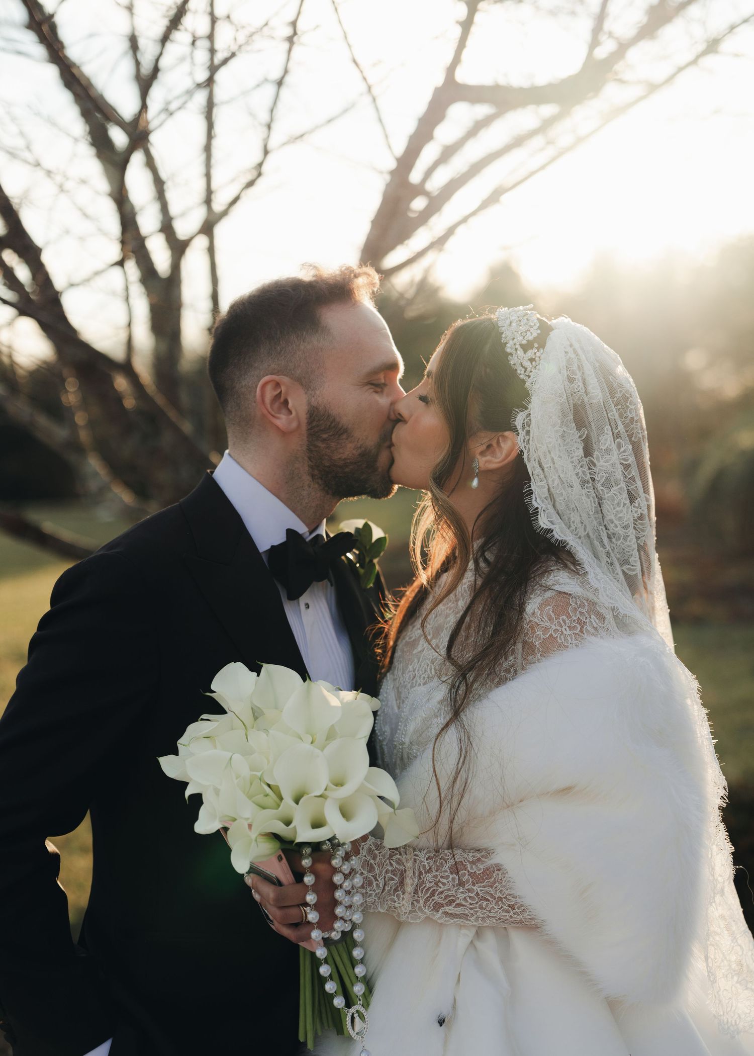 Bride and groom share a romantic kiss at golden hour, bride holding white calla lilies and rosary.