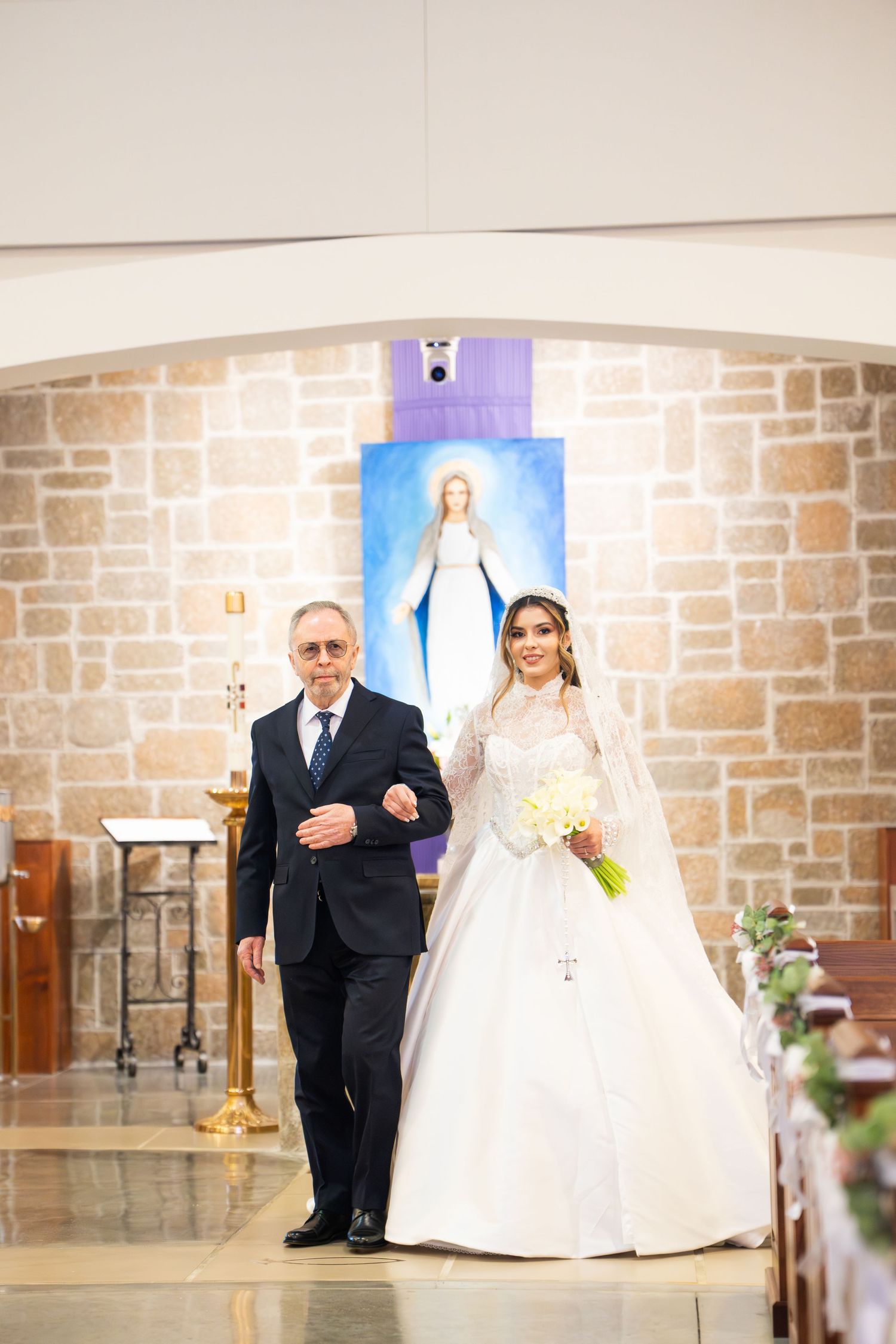 Father escorts bride in white ball gown with veil down church aisle past Virgin Mary painting.