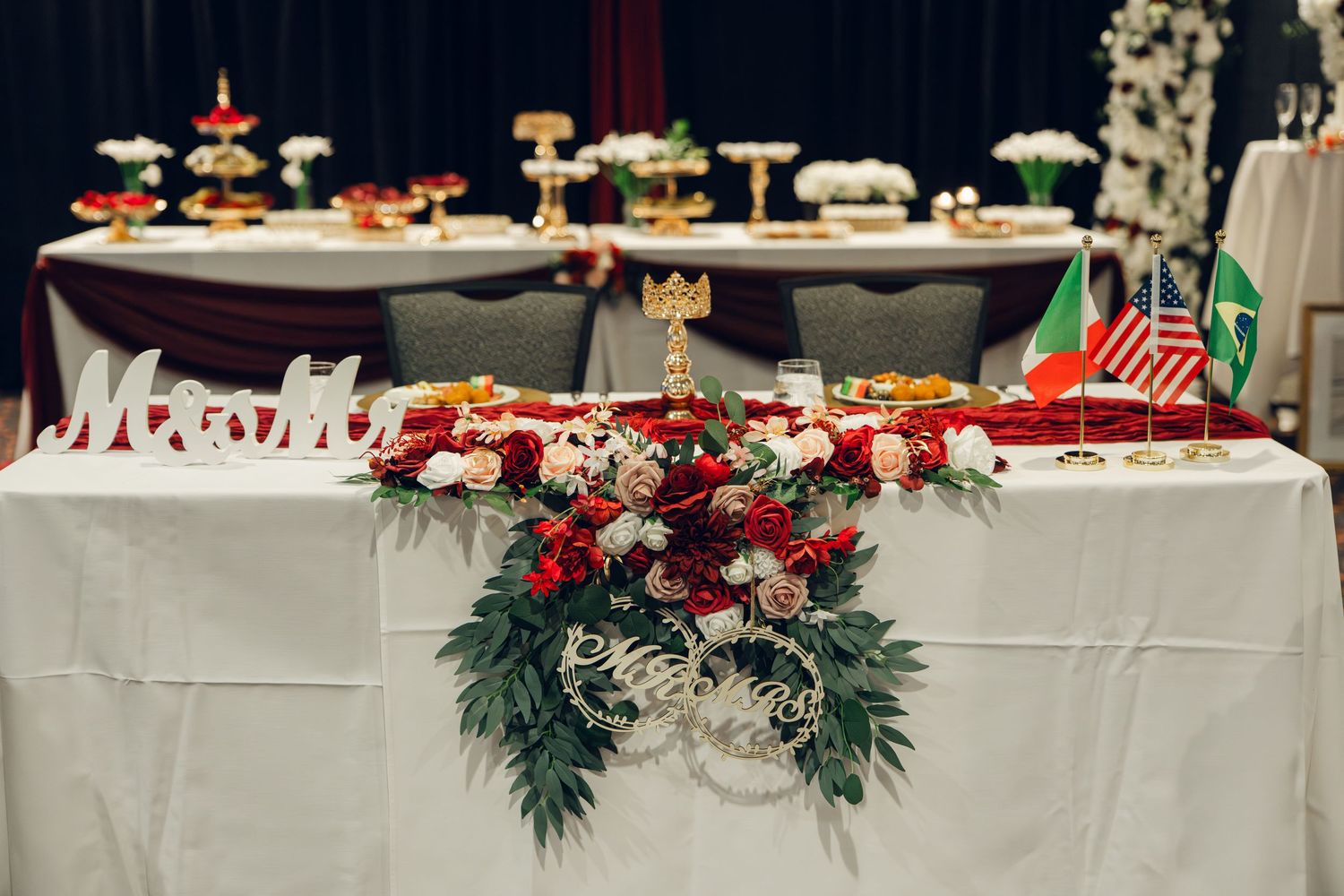 Wedding sweetheart table decorated with lush red and white floral garland, greenery, and international flags.