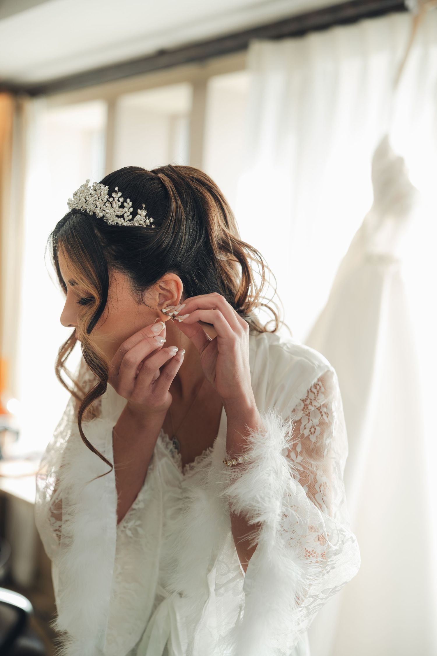 Bride in lace robe putting on earrings with wedding dress hanging in background.